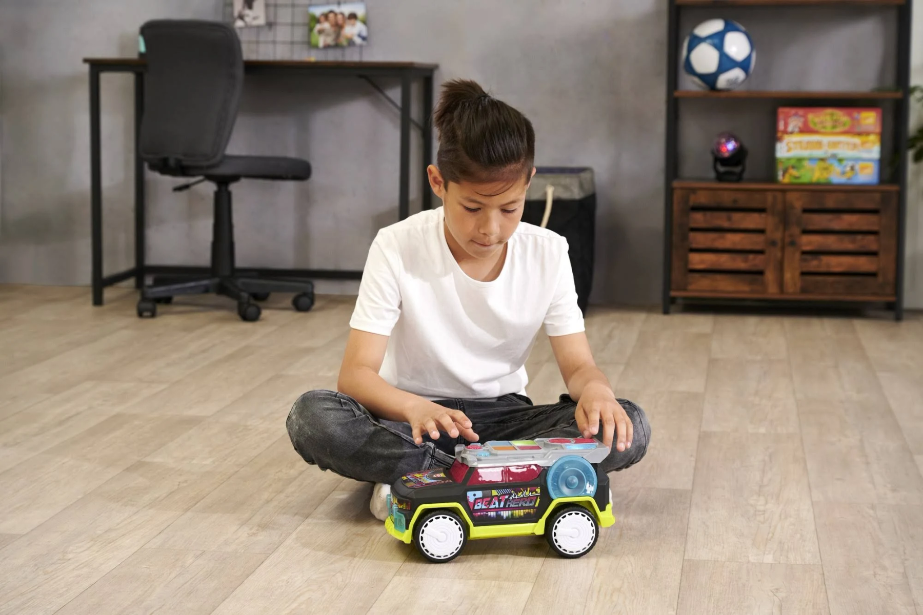 Chłopiec siedzi na podłodze z zabawką samochodem. Samochód jest kolorowy, ma światła. Tło to pokój z półkami i krzesłem.
