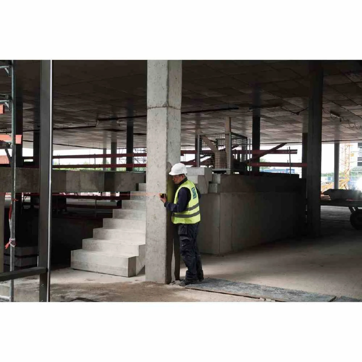 Ein Arbeiter in einer Weste misst eine Betonsäule auf einer Baustelle. Treppen und ein Gebäude sind im Hintergrund.