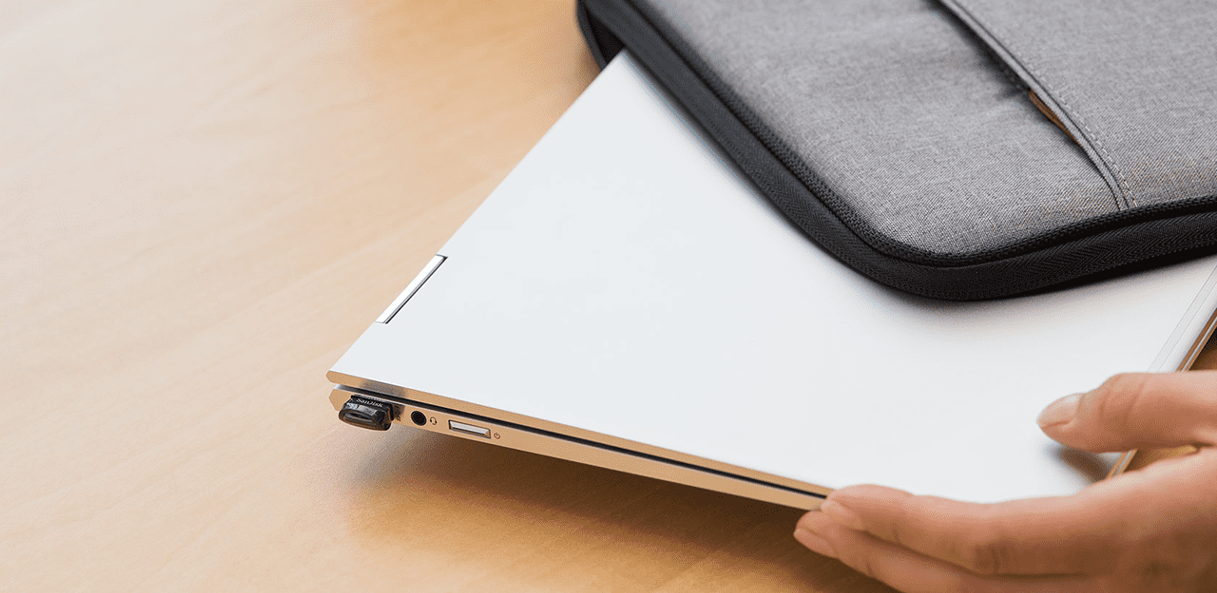 Srebrny laptop w szarej obudowie, z podłączonym donglem USB-C. Na jasnej drewnianej powierzchni.