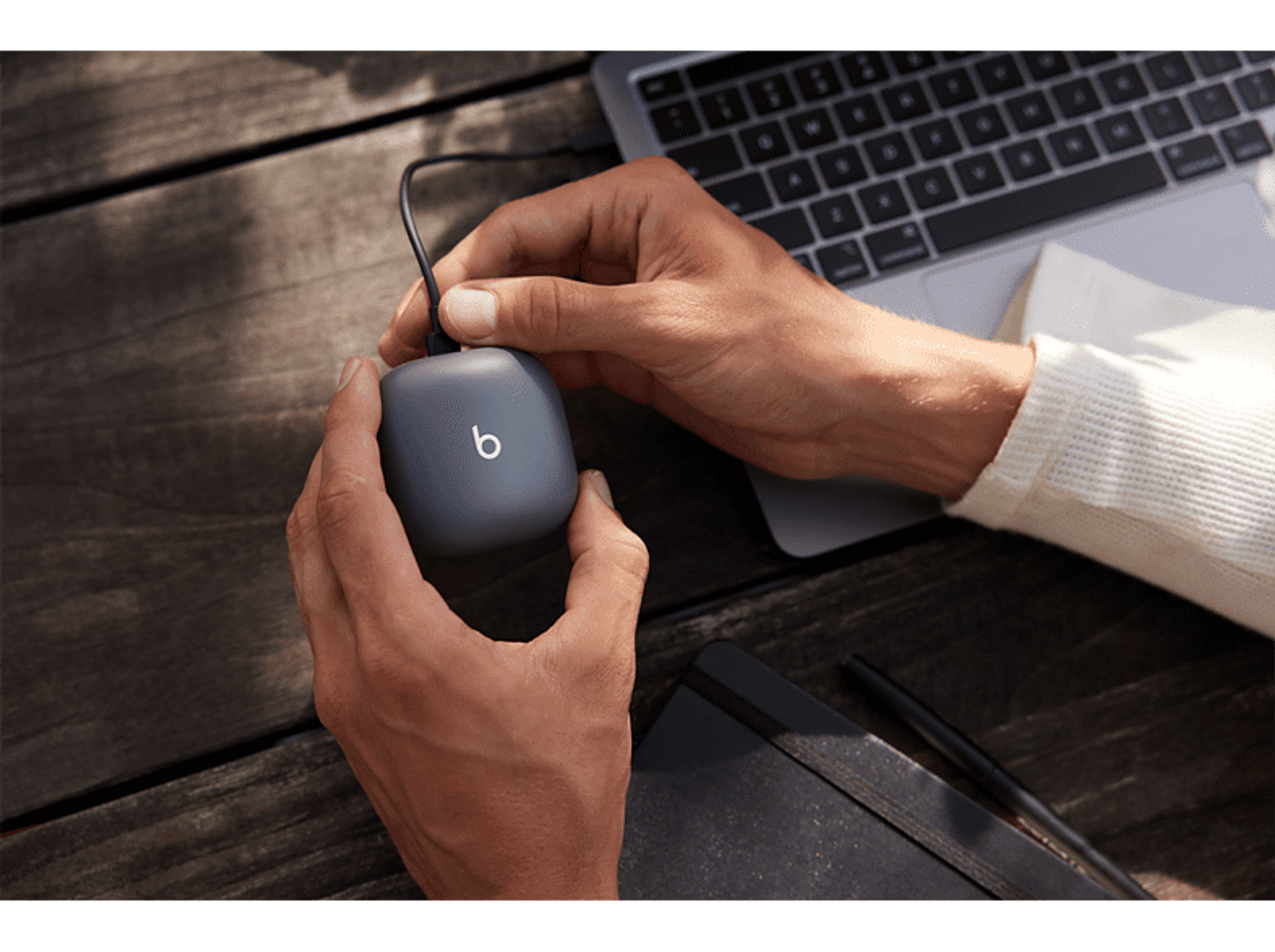 Ręce trzymające szare urządzenie ładujące z logo 'b', obok laptopa, na drewnianej powierzchni.