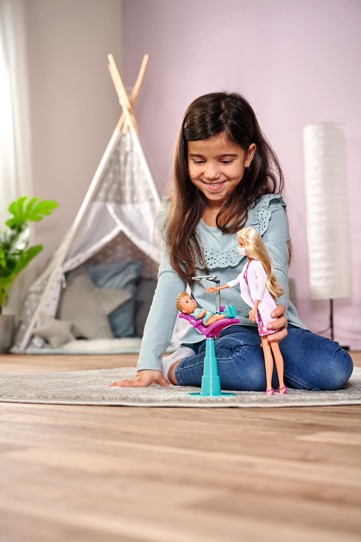 Ein junges Mädchen lächelt, während es mit einer Puppe spielt, die einen Zahnarzt nachahmt. Ein Puppenstuhl und ein Zelt sind sichtbar.