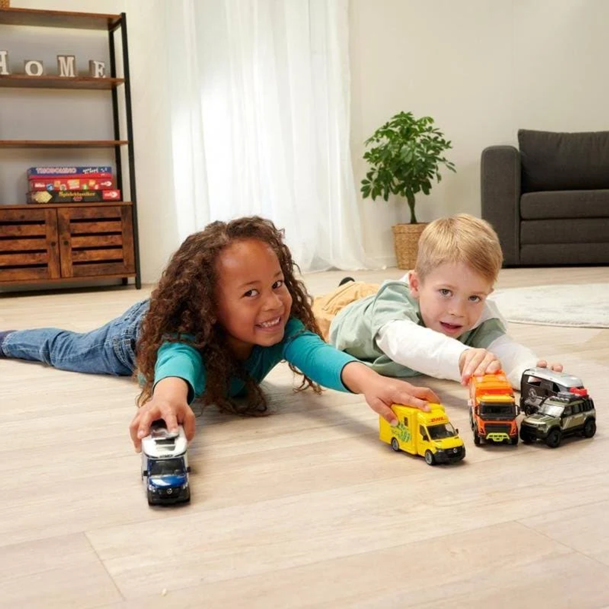Zwei Kinder spielen mit Spielzeug-LKWs auf dem Boden. Holzschrank und Zimmerpflanze im Hintergrund.