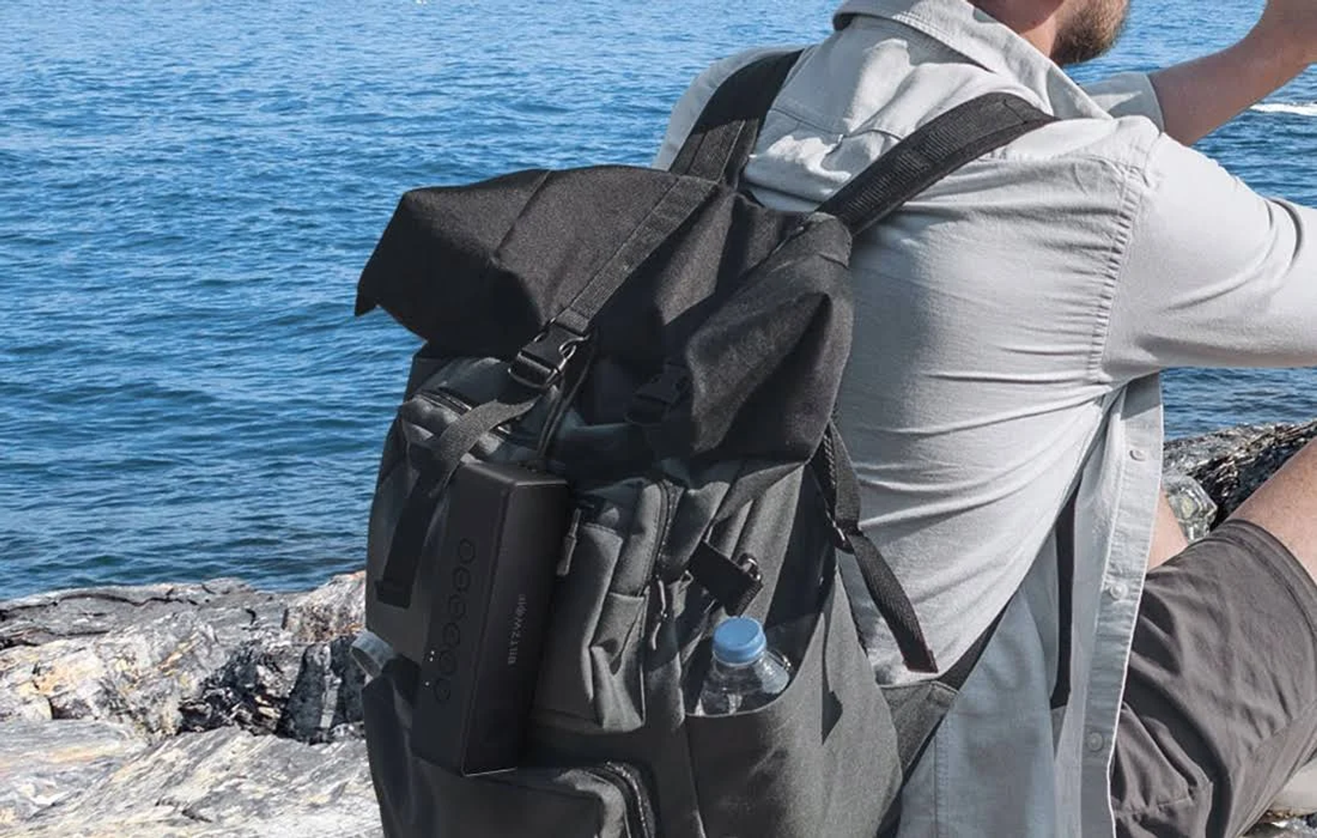 Osoba z plecakiem siedzi nad morzem. Głośnik jest przymocowany do plecaka.
