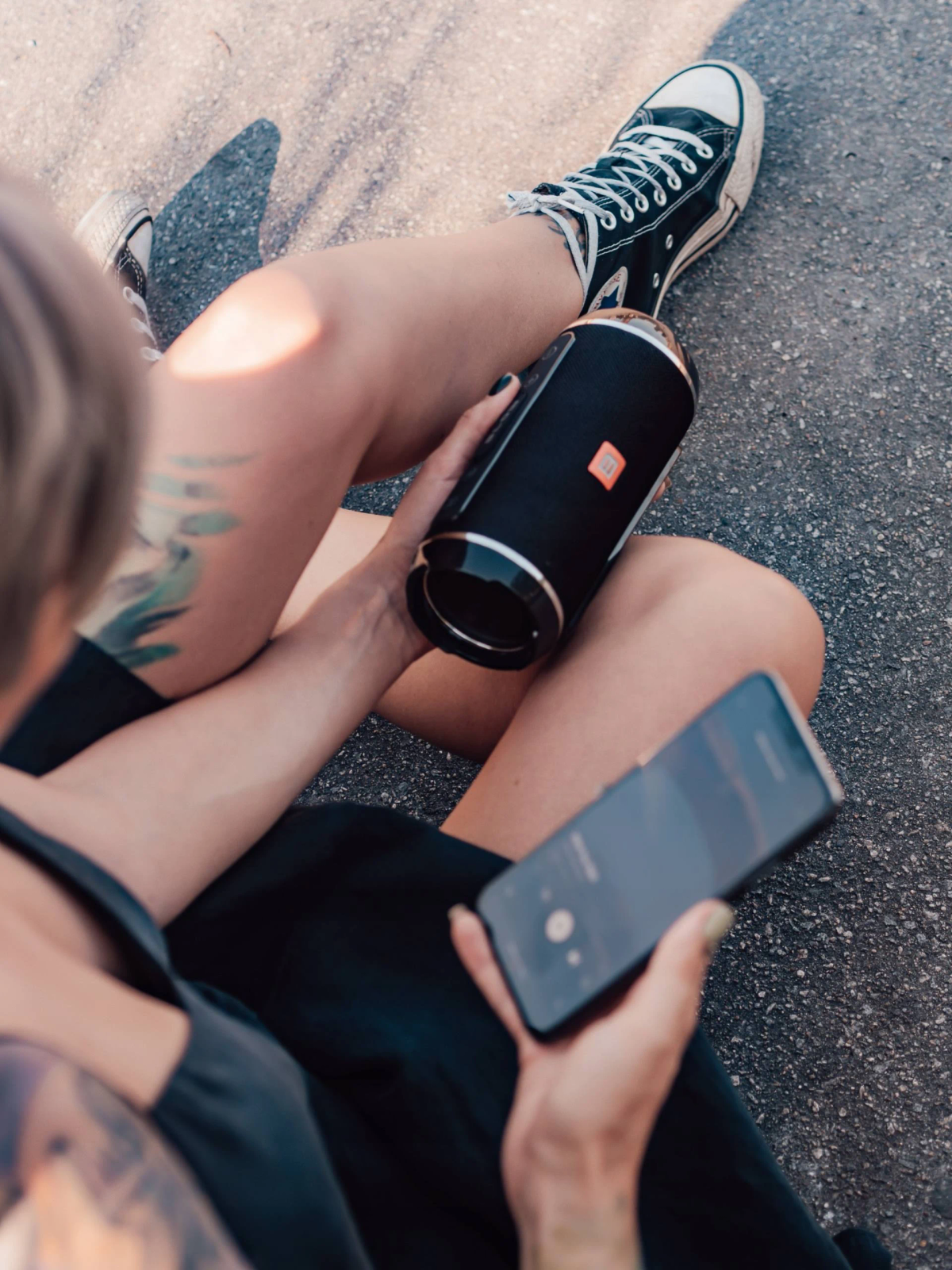 Osoba siedzi trzymając czarny głośnik i telefon. Widoczne czarne buty i szorty.