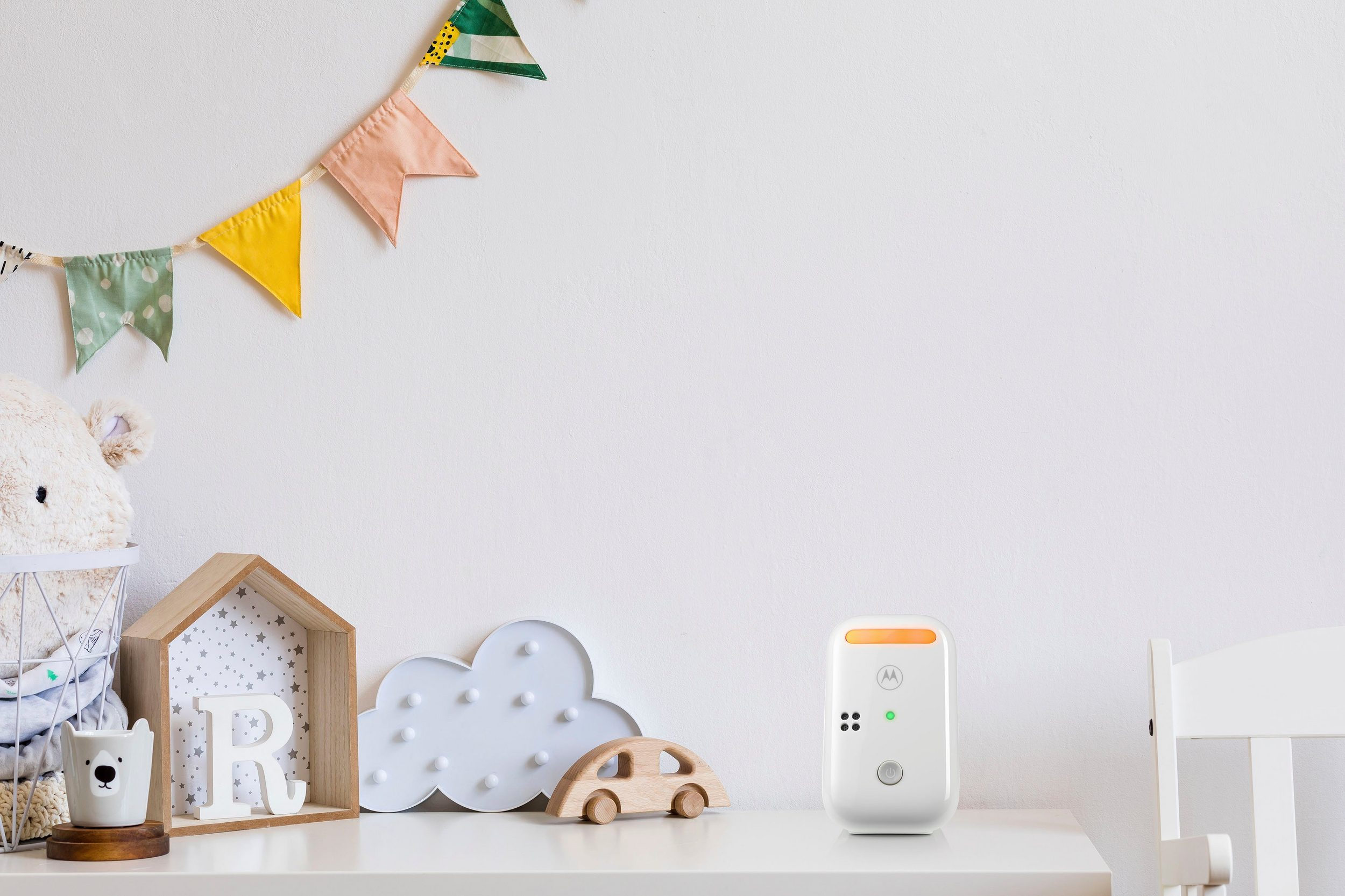 Ein Babyphone auf einem weißen Tisch, mit einer dekorierten Wand und Spielzeug im Hintergrund.