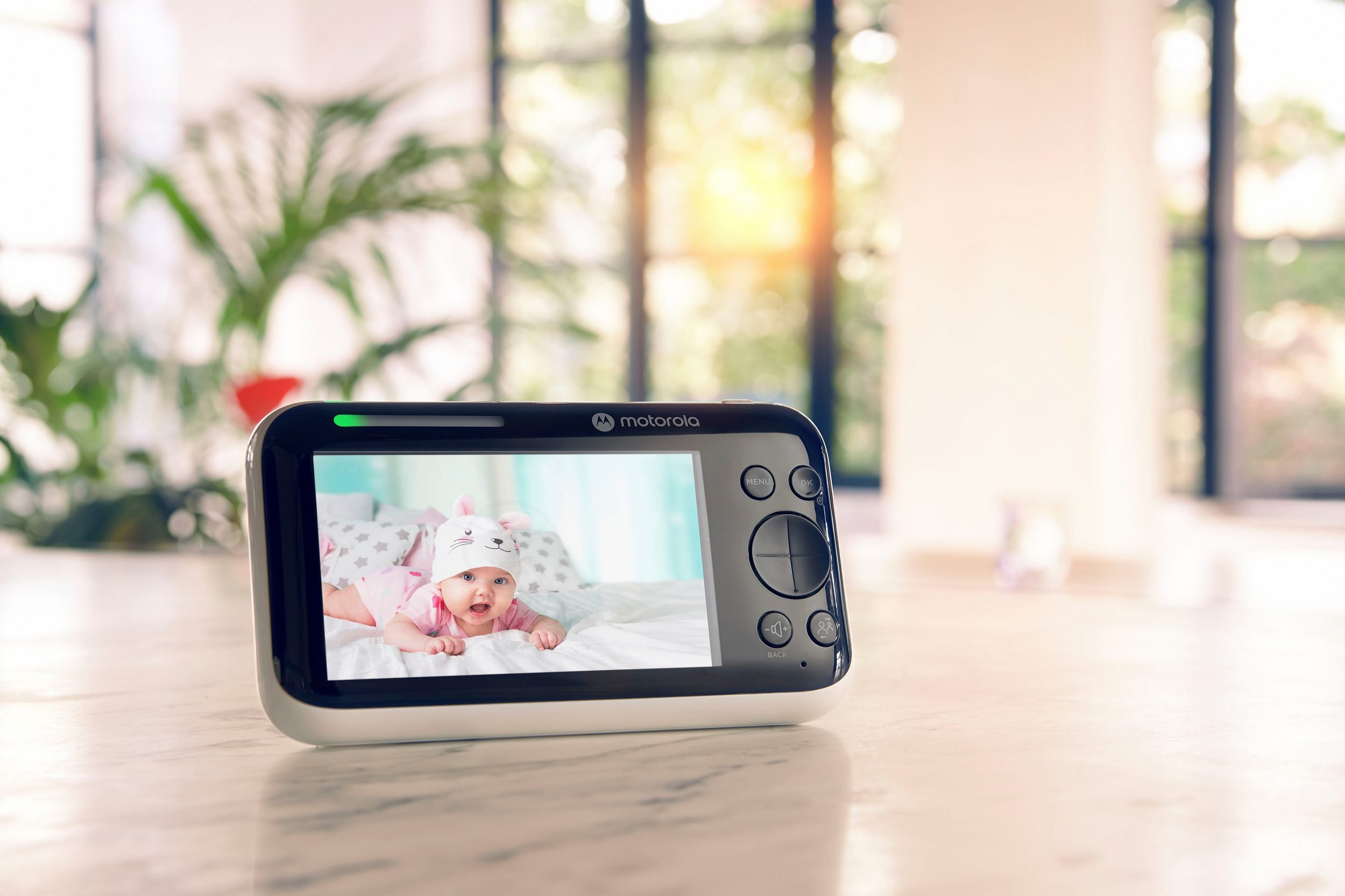 Babymonitor met een baby op het scherm, liggend op een bed, met een hoedje, binnenshuis.