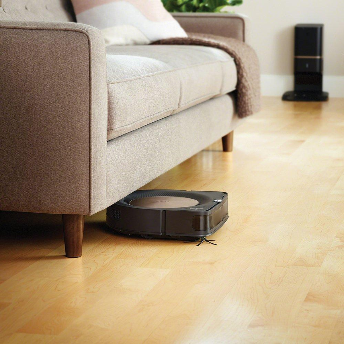 Ein Roboter-Staubsauger unter einer beigen Couch auf hellem Holzboden. Ein hoher schwarzer Turm im Hintergrund.