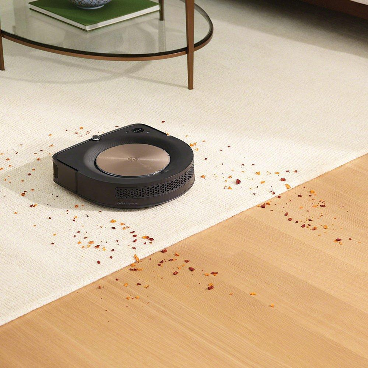 Schwarzer Roboterstaubsauger auf einem weißen Teppich mit Ablagerungen, in der Nähe eines Tisches und eines Holzbodens.