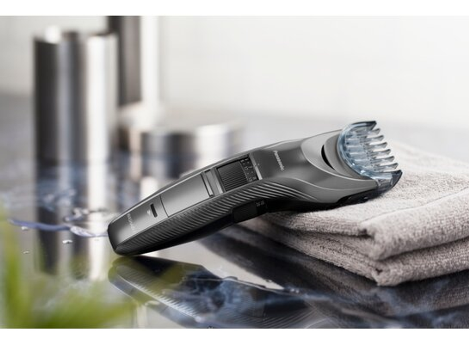 Un rasoio elettrico grigio scuro su un asciugamano, con due contenitori metallici dietro.