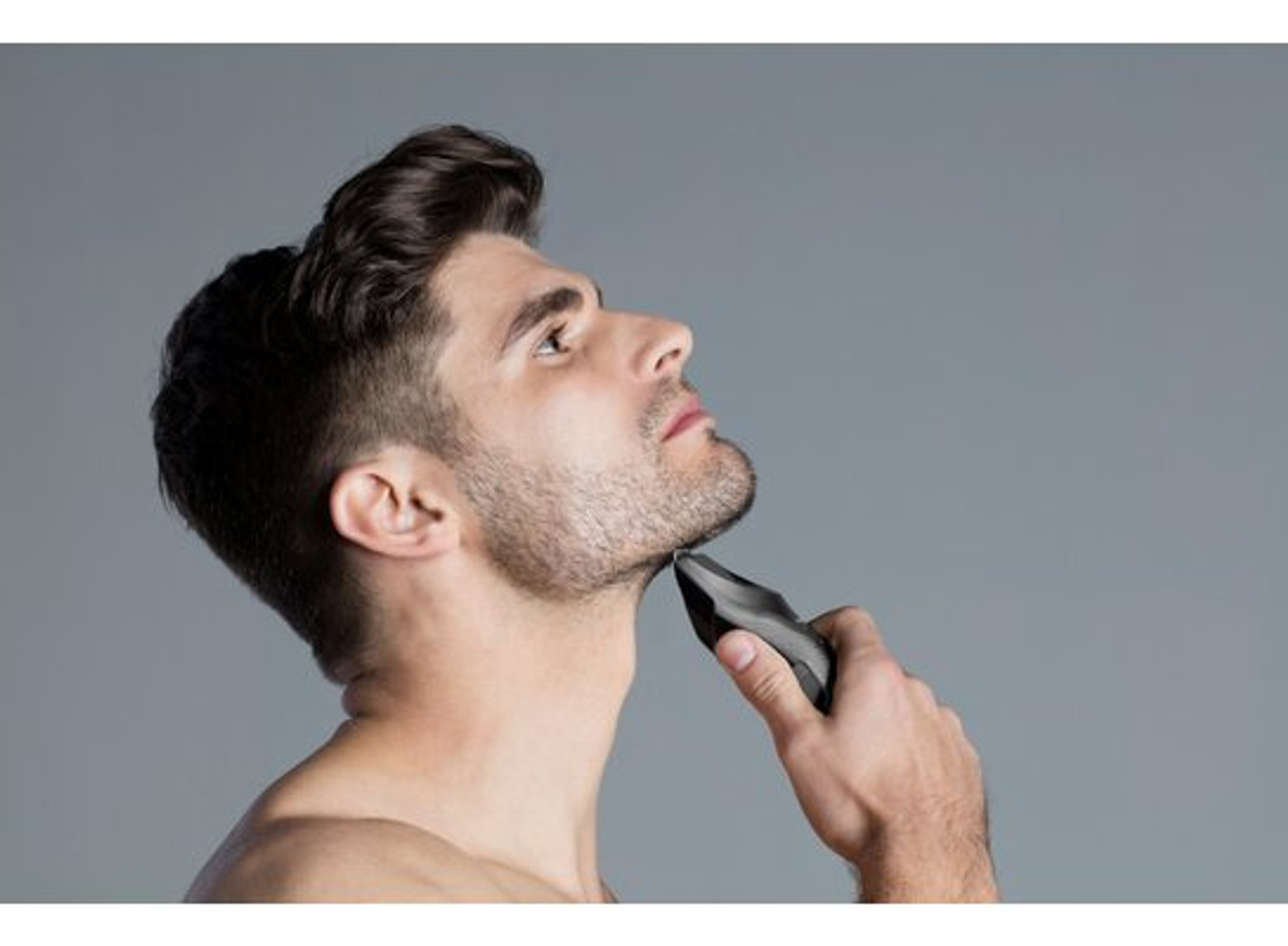 Un uomo si taglia la barba con un regolabarba elettrico, guardando in alto.