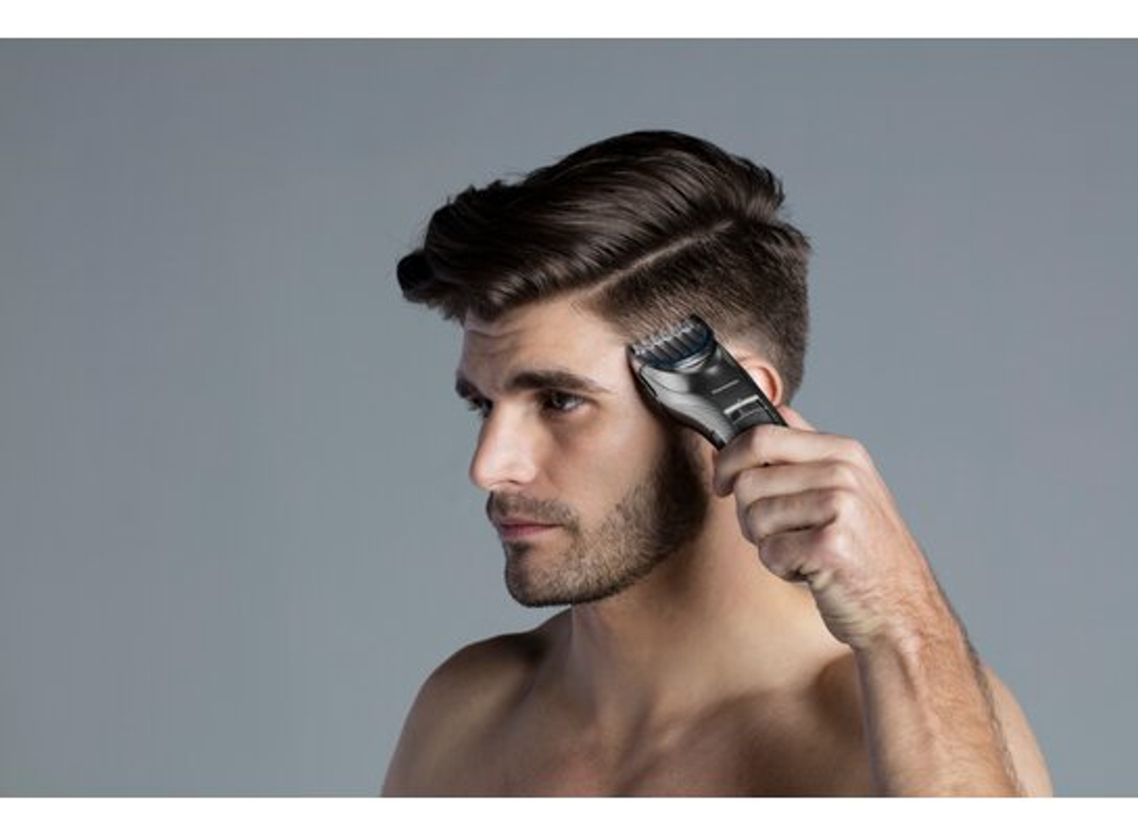 Uomo che si taglia i capelli con un tagliacapelli nero, vista laterale, sfondo grigio.