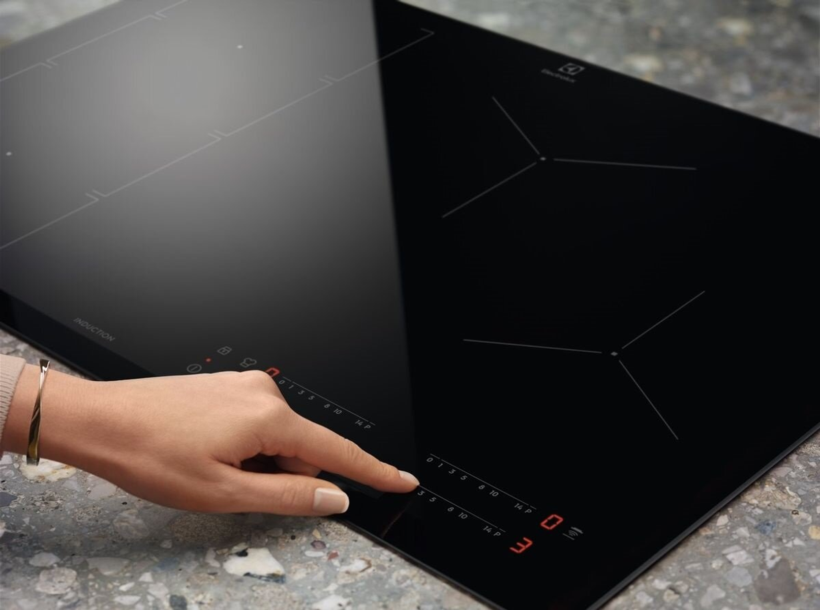 Ręka dotyka czarnej płyty indukcyjnej. Widoczne elementy sterujące na dole. Logo Electrolux u góry po prawej.