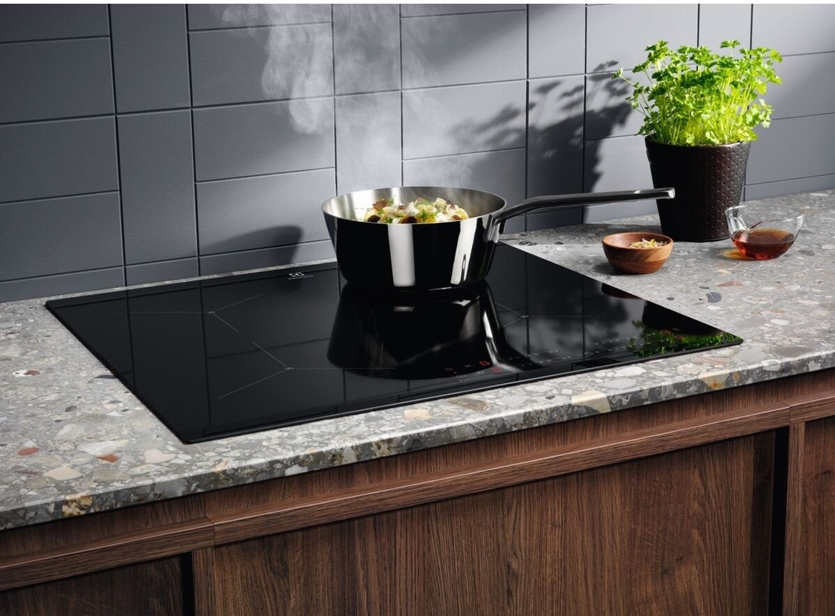 Czarna płyta indukcyjna z patelnią gotującą jedzenie. Szara kafelkowa ściana i roślina w doniczce.