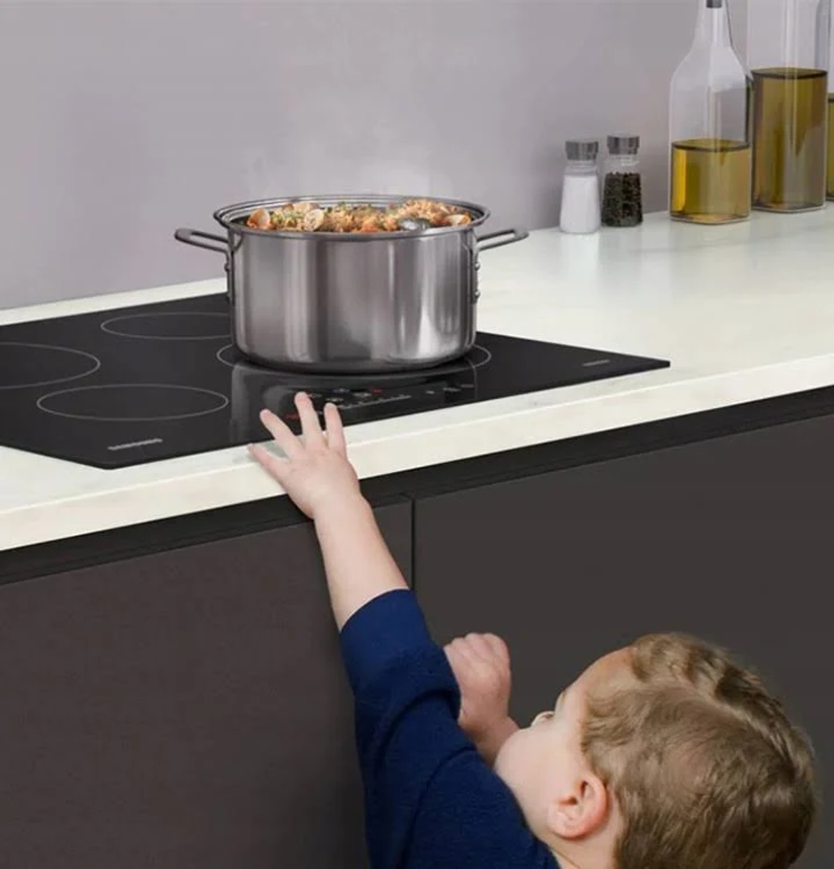 Un niño alcanza una olla en una estufa. La olla tiene tapa, la estufa es negra.