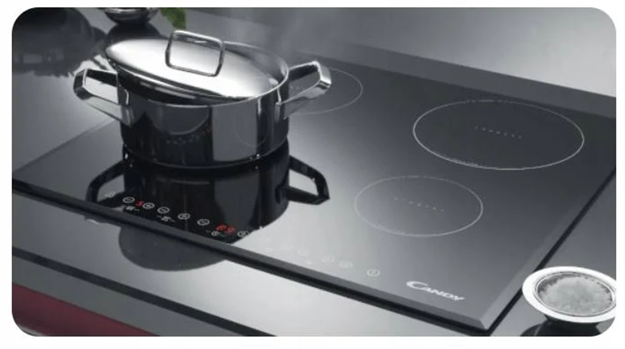 Czarna płyta indukcyjna z garnkiem i trzema nieużywanymi palnikami. Srebrny garnek po lewej.