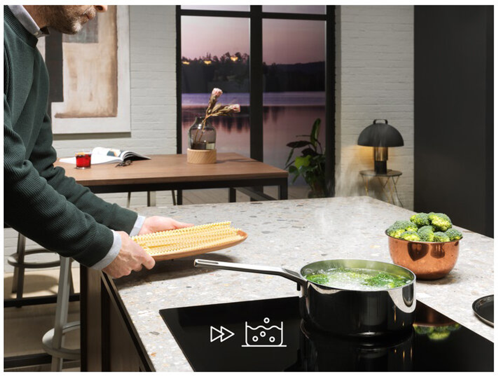 Osoba umieszczająca makaron na blacie, z garnkiem wrzącym na czarnej płycie indukcyjnej. Tło kuchni.