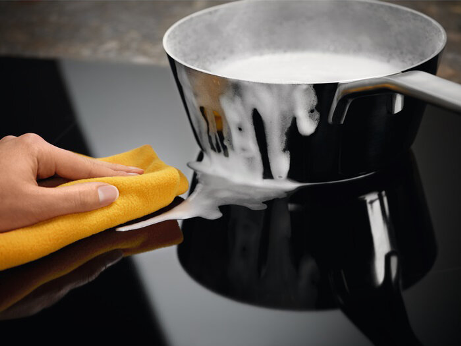 Ręka czyszcząca czarny garnek żółtą szmatką na czarnej płycie indukcyjnej.