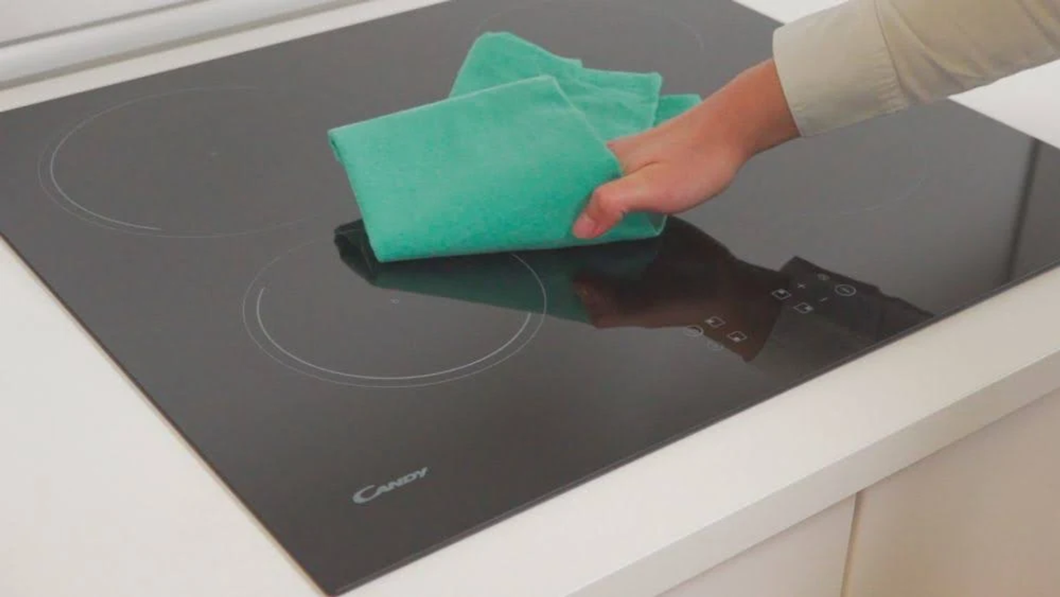 Dłoń wycierająca czarną płytę grzejną zieloną szmatką. Biały blat w tle.