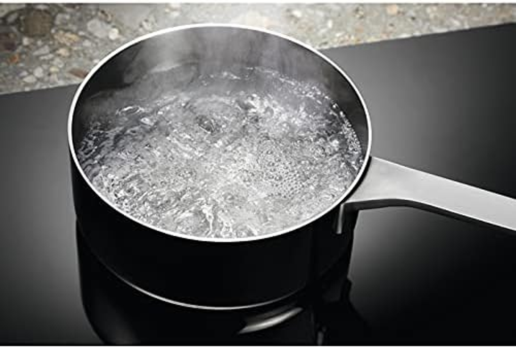 Garnek z wrzącą wodą na czarnej płycie kuchennej. Para unosi się z wody.