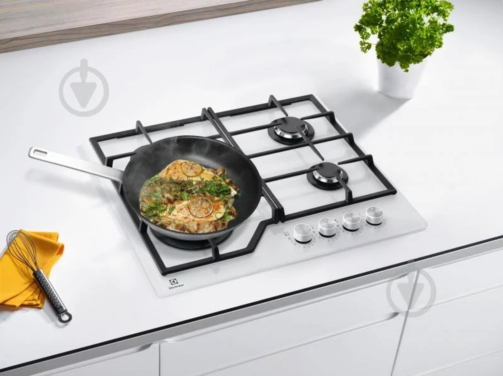 Biała płyta kuchenna z czarną ramą, patelnia z gotowanym jedzeniem i roślina w tle.