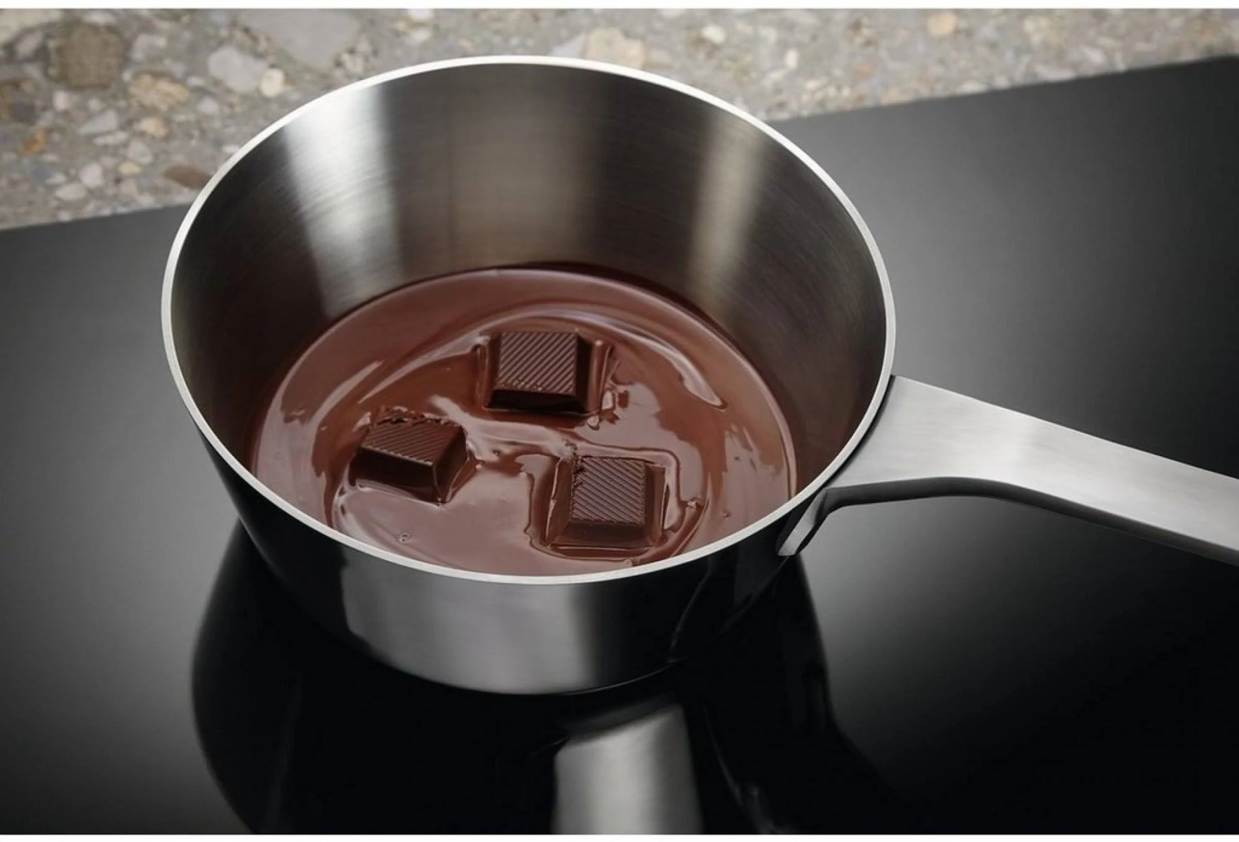 Una pentola con cioccolato fuso e quadratini di cioccolato su un piano cottura nero.