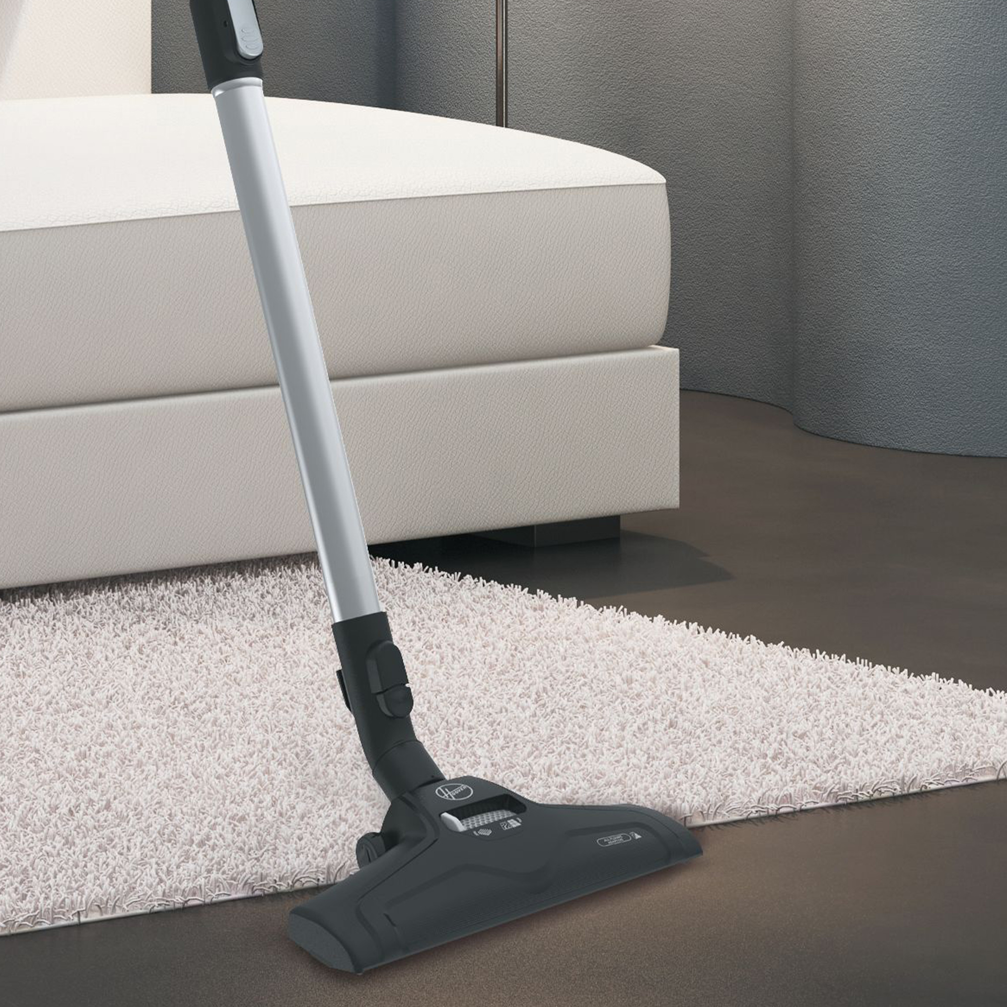 Testa dell'aspirapolvere su un tappeto bianco. L'aspirapolvere è grigio scuro, con un lungo manico argentato.
