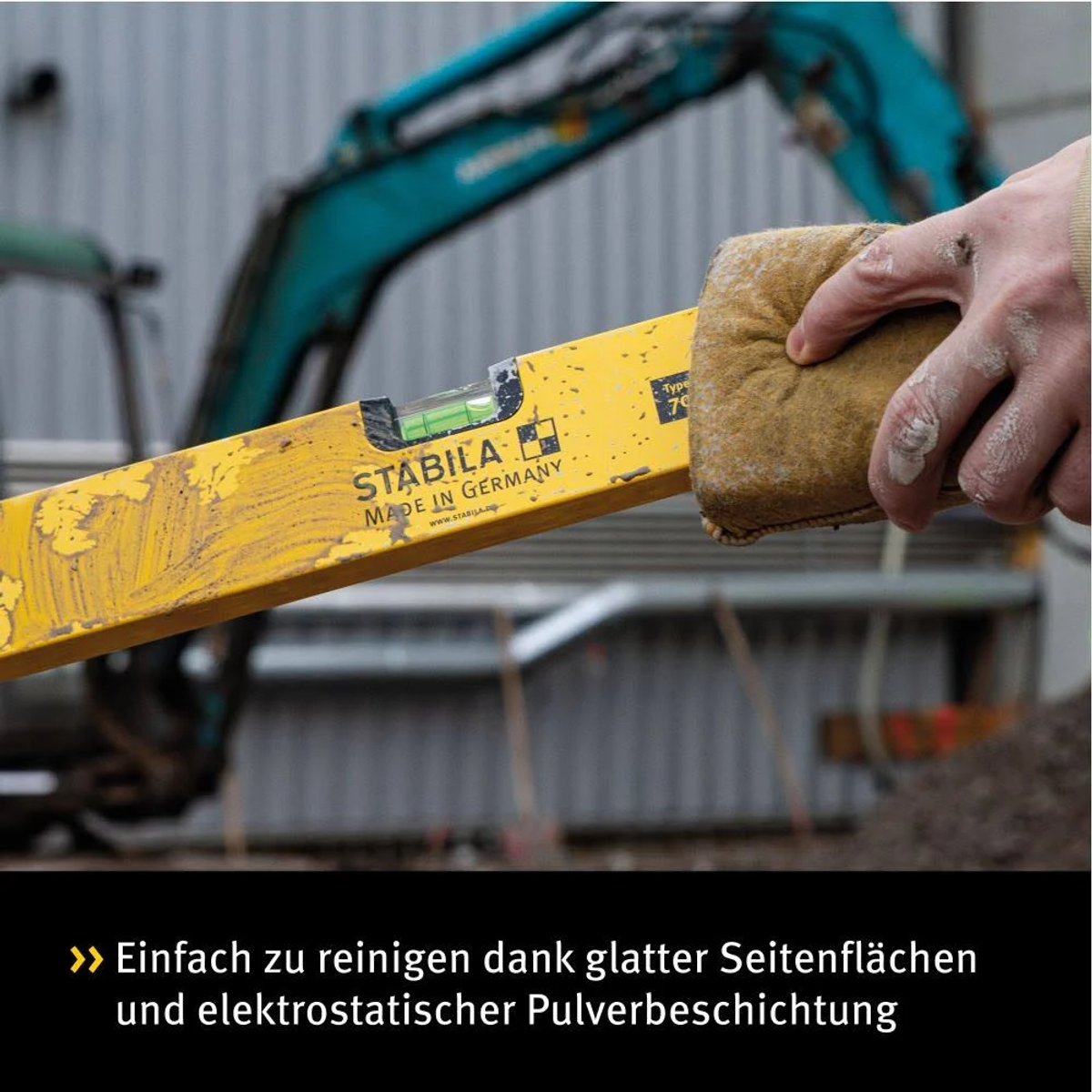 Eine gelbe Wasserwaage wird mit einem braunen Tuch von einer Hand gereinigt. Baustelle im Hintergrund.