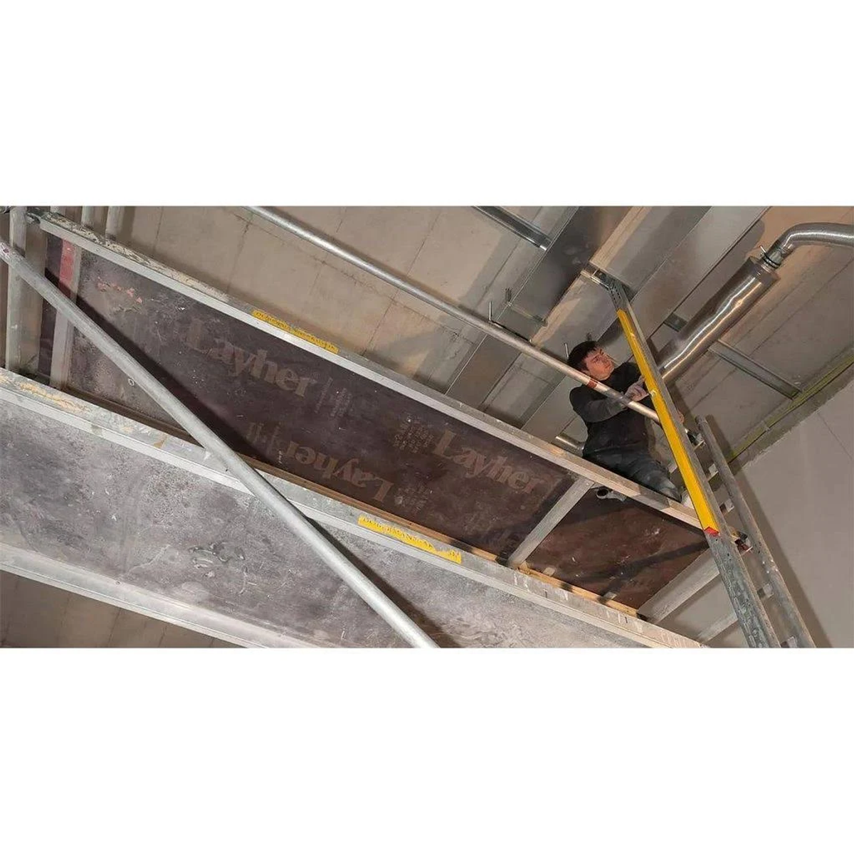 Ein Arbeiter auf einem Gerüst in einem Gebäudeinneren. Er arbeitet in der Nähe der Decke und Rohre.