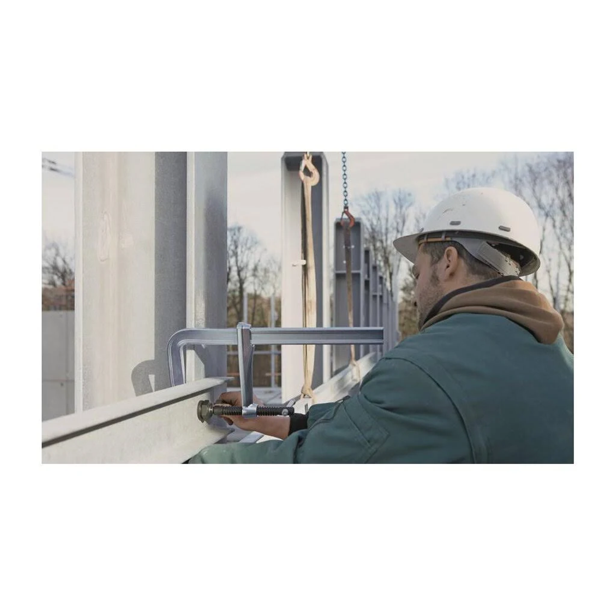 Eine Person mit Schutzhelm verwendet eine Klammer, um einen Metallträger an einer größeren Struktur zu befestigen.
