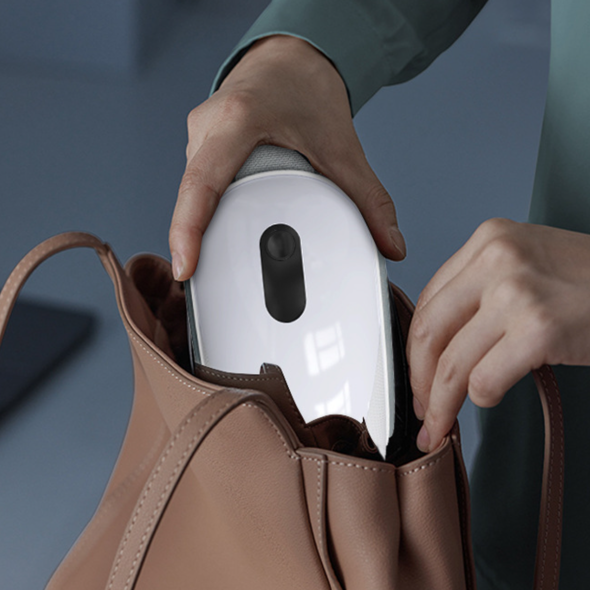 Hände, die ein weißes Gerät in eine hellbraune Handtasche stecken.