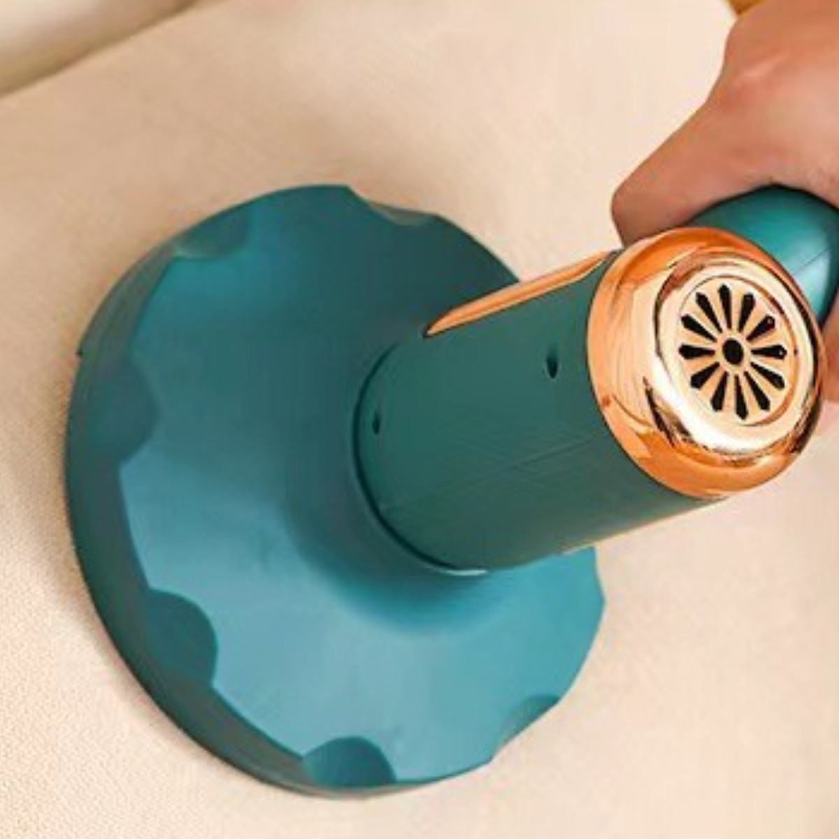 Ein teal-farbener und kupferfarbener Staubsauger liegt auf einem beigen Kissen. Eine Hand hält den Griff.