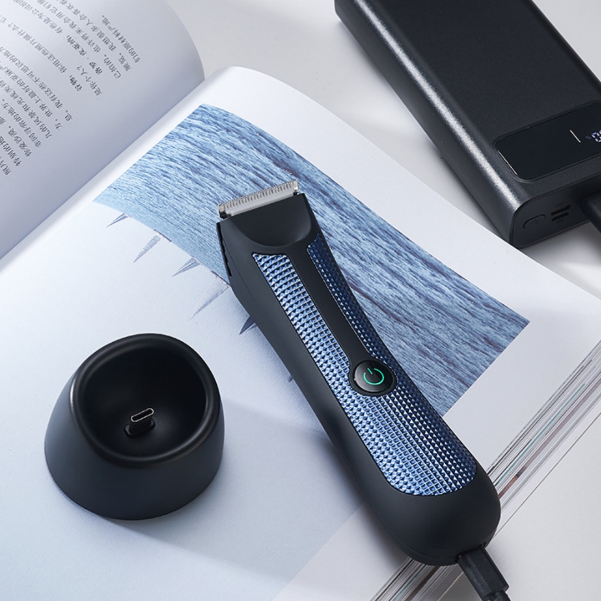 Ein schwarzer Barttrimmer mit Ladestation, Powerbank und Zeitschrift im Hintergrund.