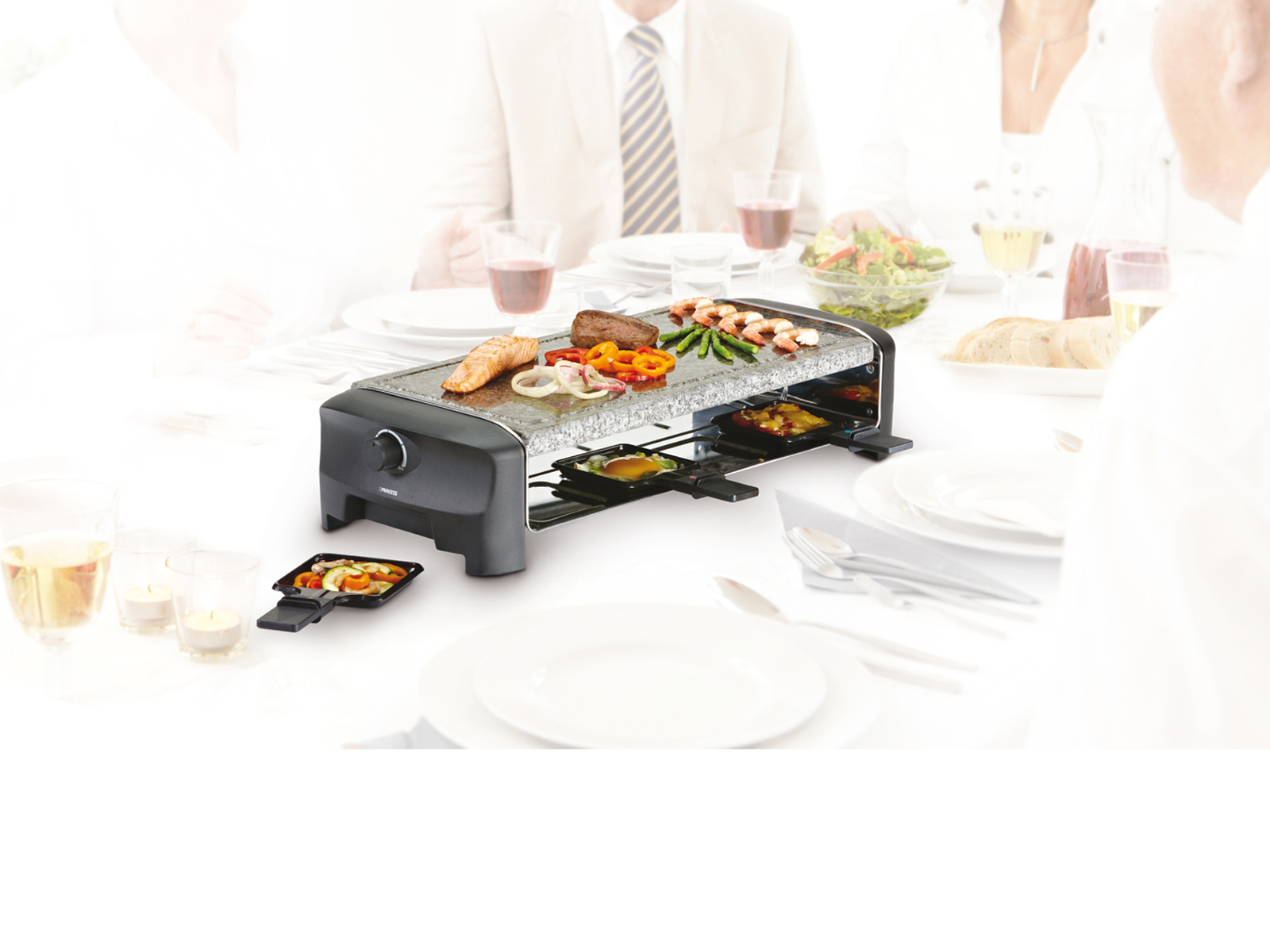Raclette-Grill mit Essen, neben einem Tisch mit Personen im Hintergrund.