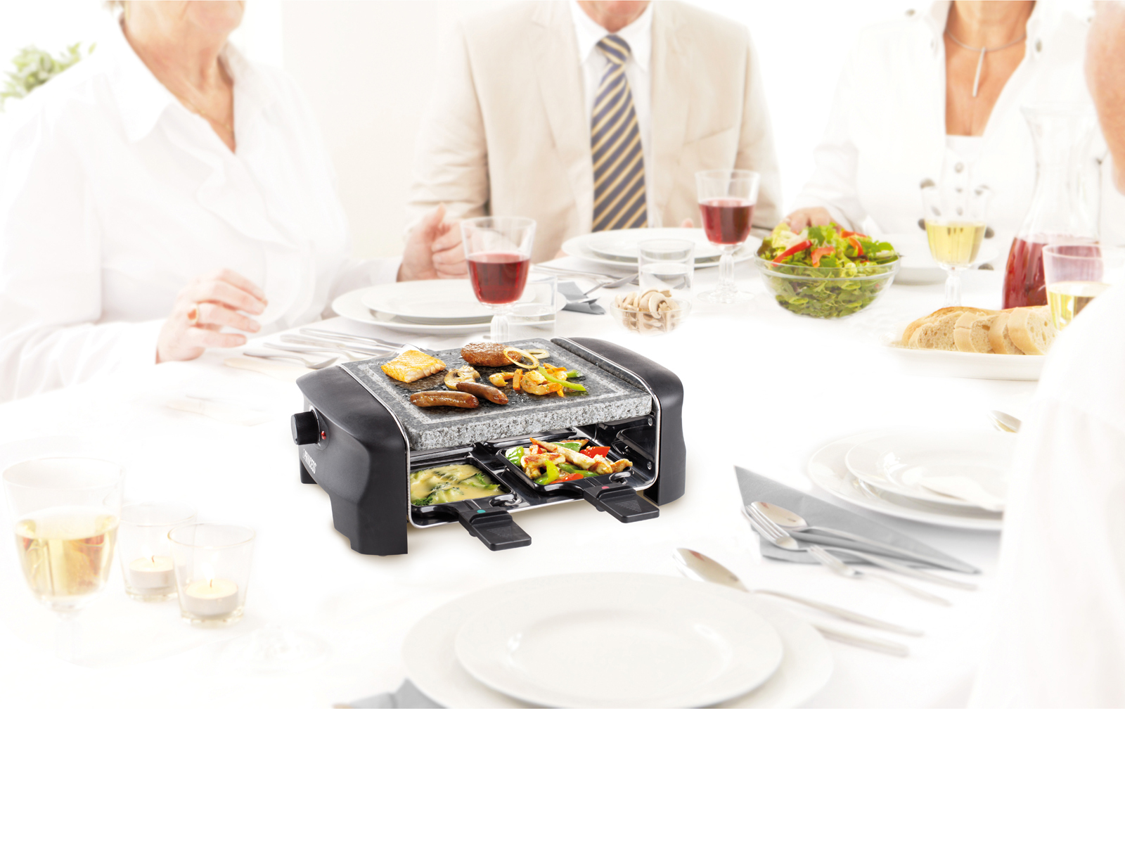 Ein Raclette-Grill mit Essen auf einem weißen Tisch. Menschen sitzen drum herum. Gläser und Teller sind vorhanden.