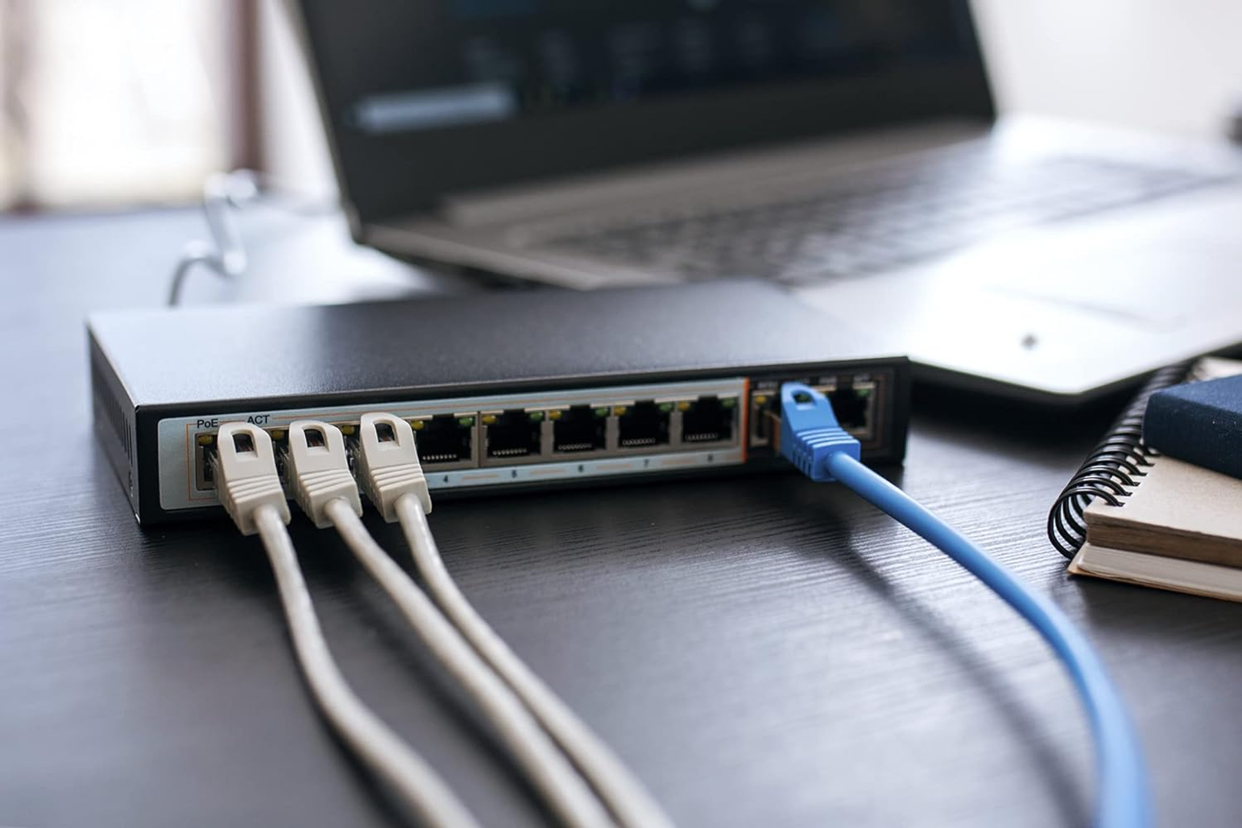 Switch di rete nero con cavi collegati. Laptop e taccuino su una scrivania di legno.