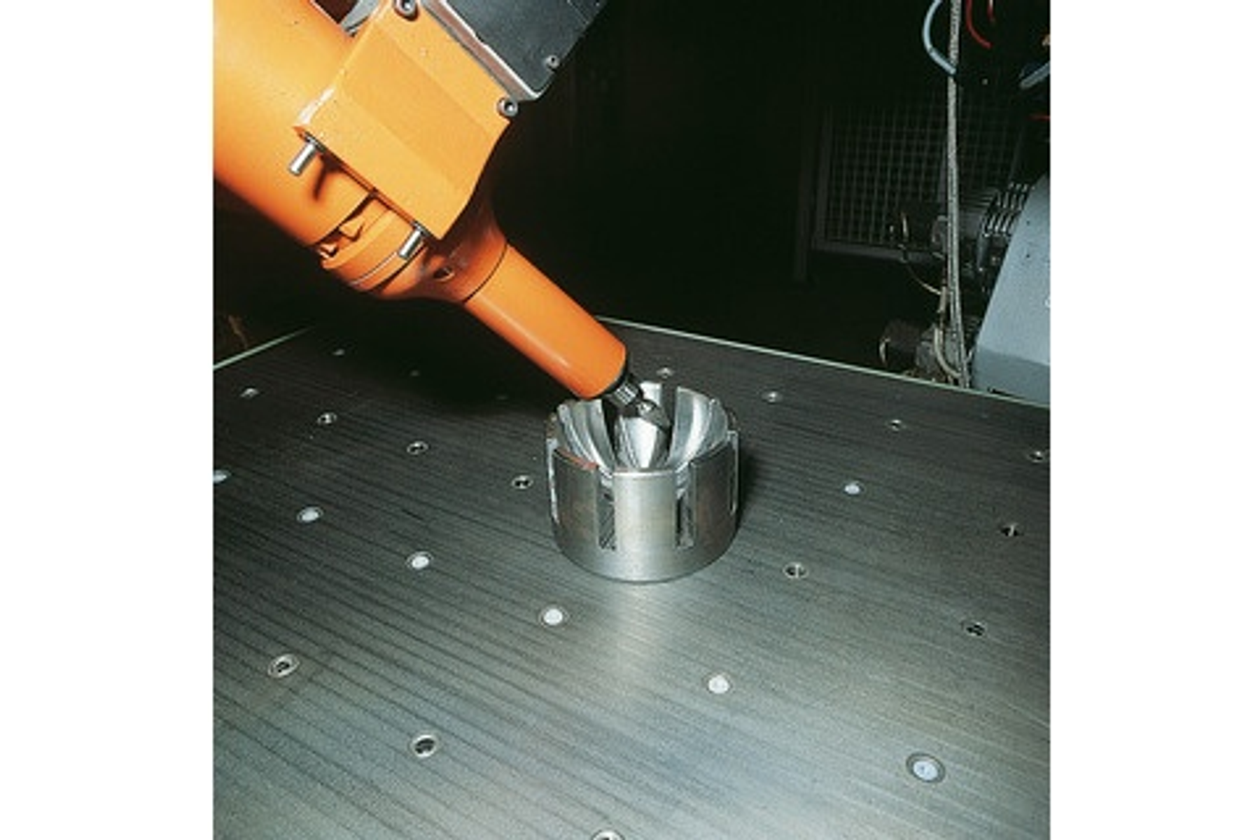 Ein Roboterarm bearbeitet ein Metallteil. Der Arm ist orange, das Teil ist silberfarben.