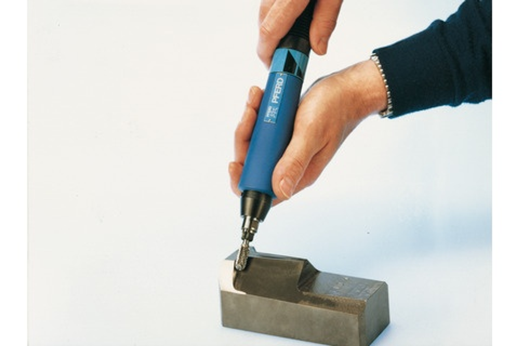 Hand hält ein blau-schwarzes Rotationswerkzeug, das einen Metallblock schleift. "PFERD" steht auf dem Werkzeug.