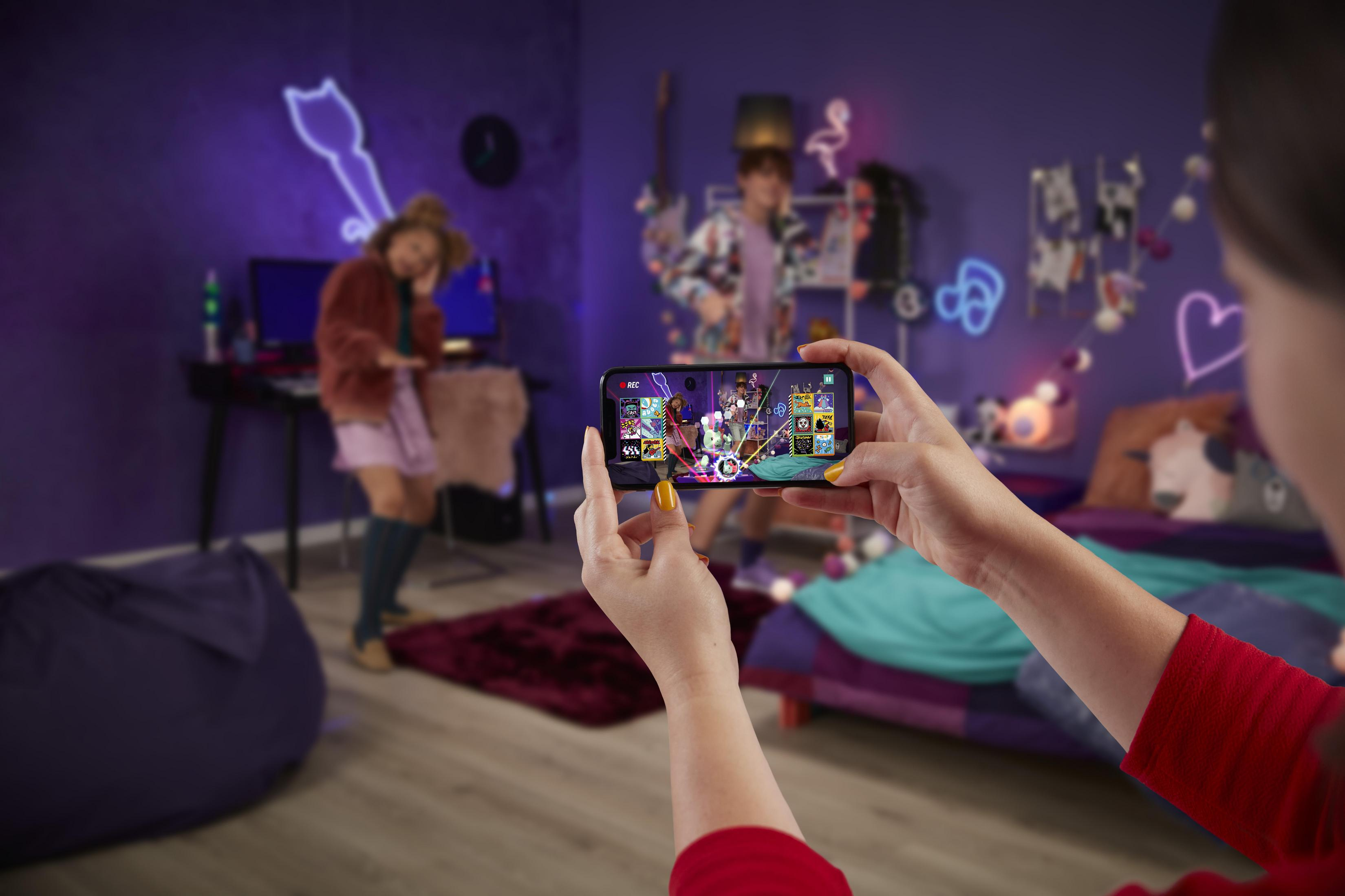 Osoba trzymająca telefon z ekranem gry. Rozmyte tło z ludźmi.