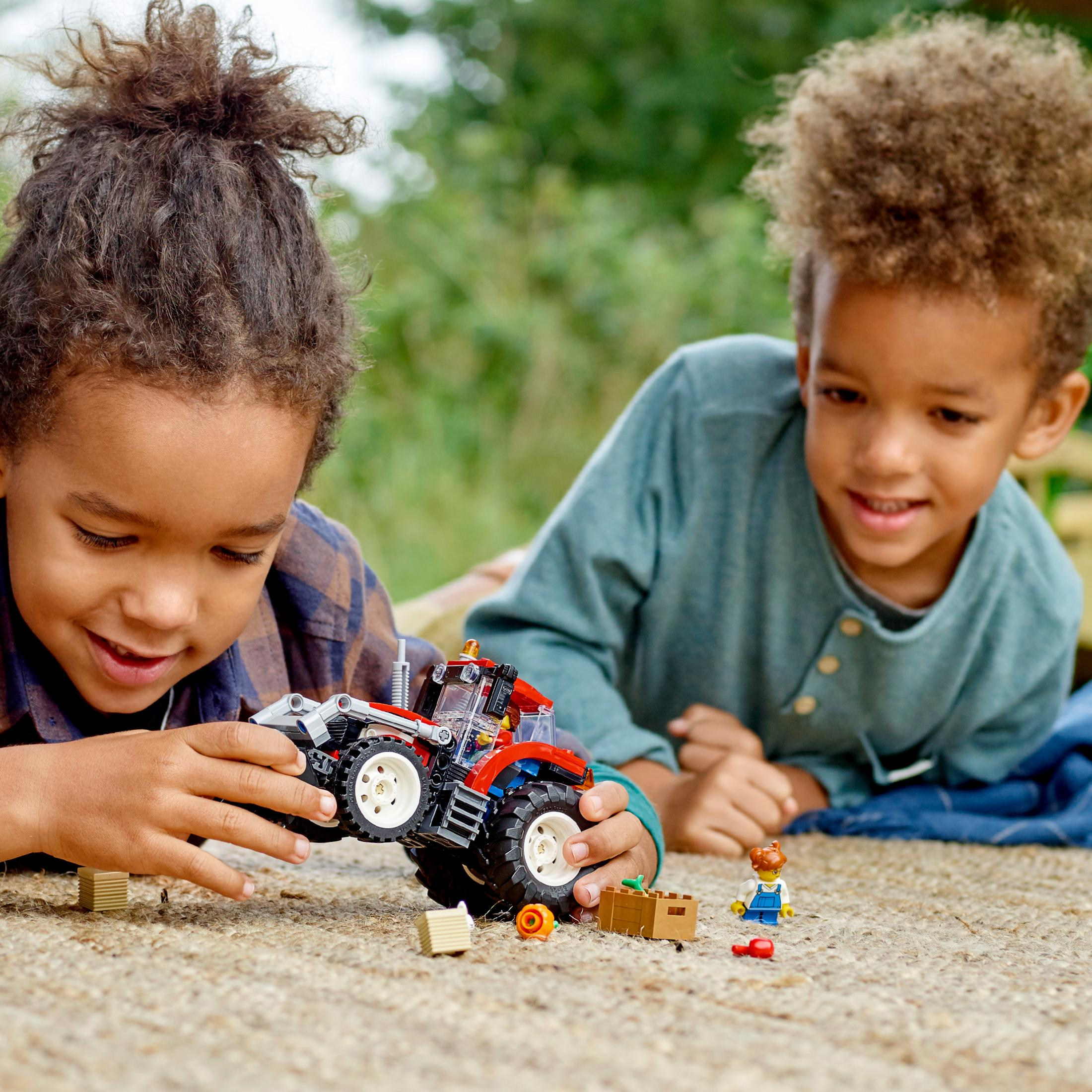 Zwei Kinder spielen mit einem roten Lego-Traktor auf einer blauen Decke, Lego-Teile herum.