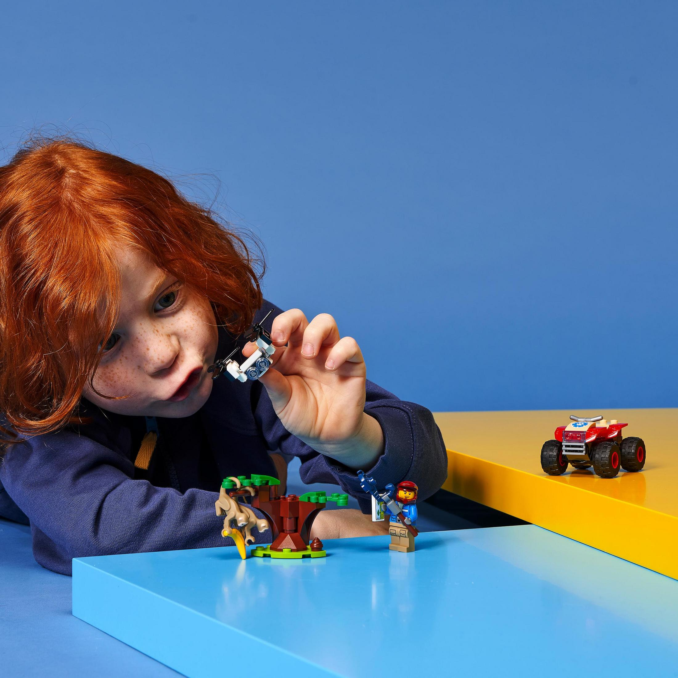 Kind spielt mit Lego-Figuren, einem roten Auto und einem Baum auf blauer und gelber Oberfläche.
