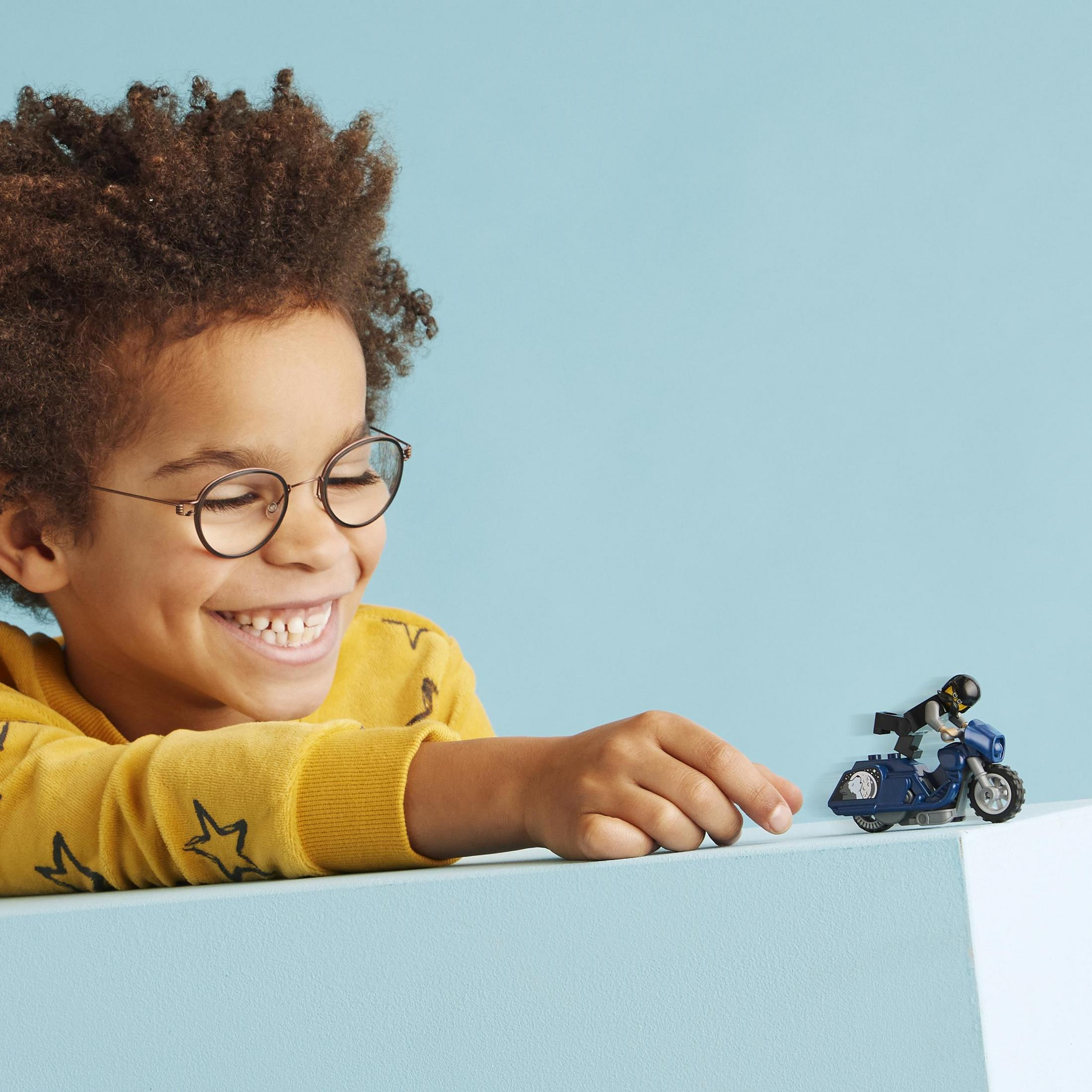 Ein Junge mit Brille lächelt einen Lego-Motorroller und eine Figur auf einer blauen Oberfläche an.