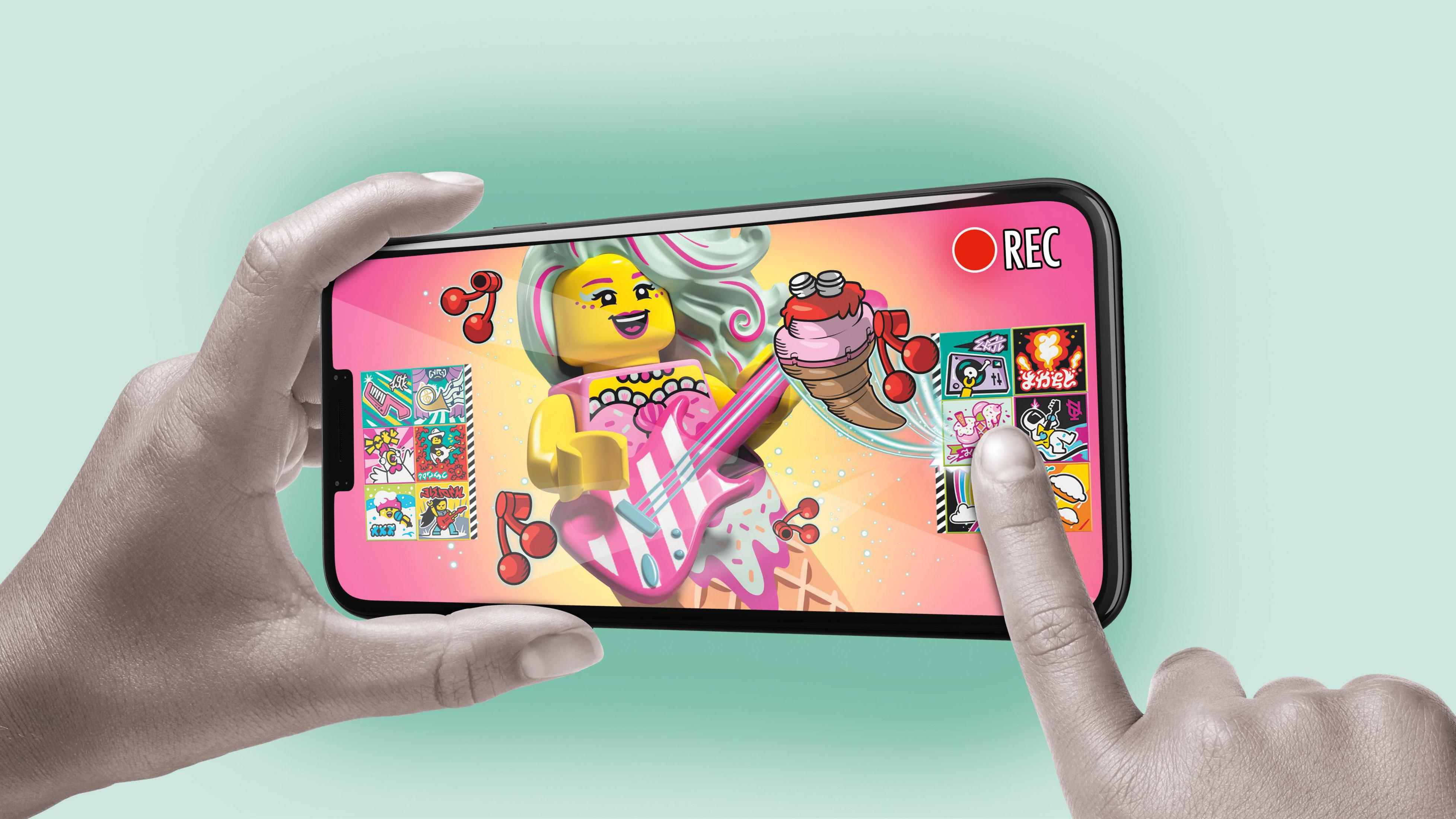 Ręka trzymająca telefon z figurką Lego grającą na gitarze na scenie.