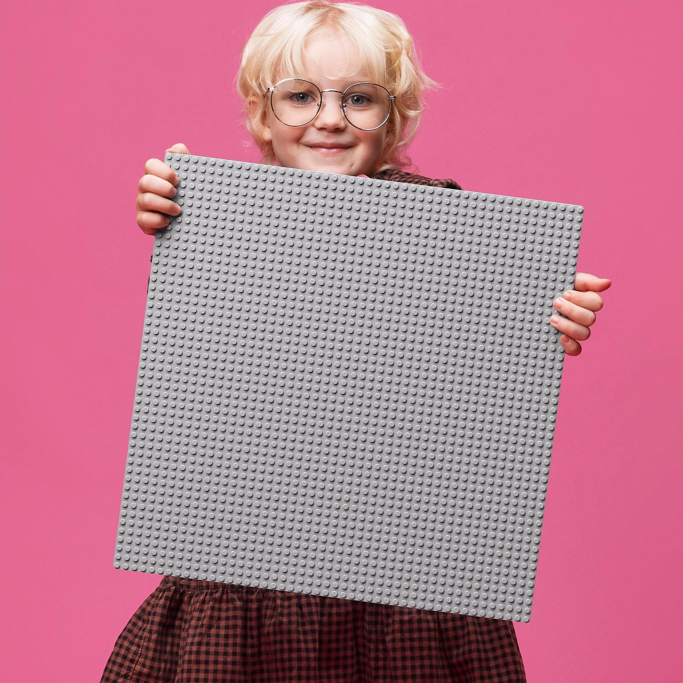 Blondynka trzymająca dużą szarą płytkę bazową Lego na różowym tle.