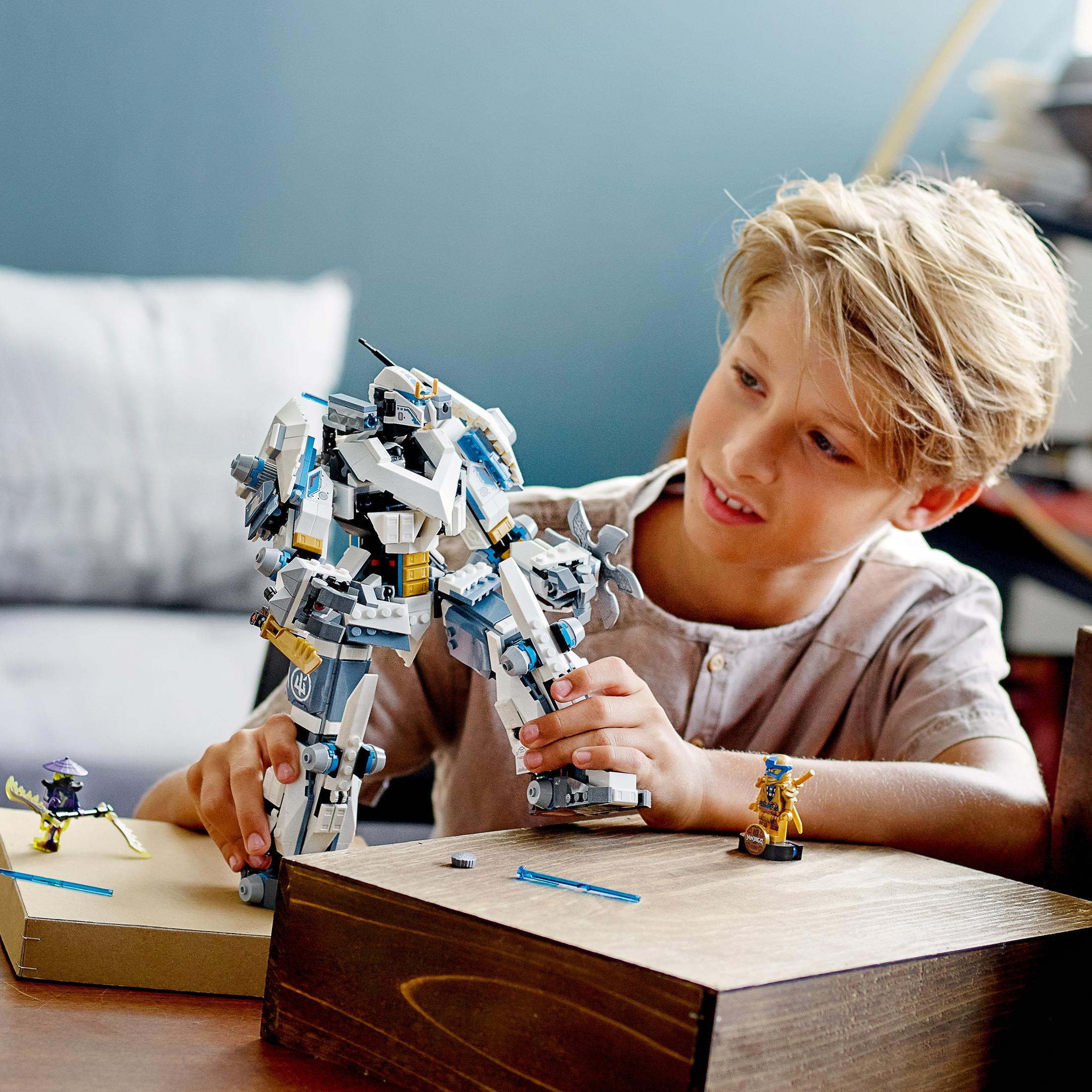 Ein Junge spielt mit einem Lego Ninjago Mech-Spielzeug. Der Junge lächelt, und der Hintergrund ist verschwommen und zeigt einen Raum.