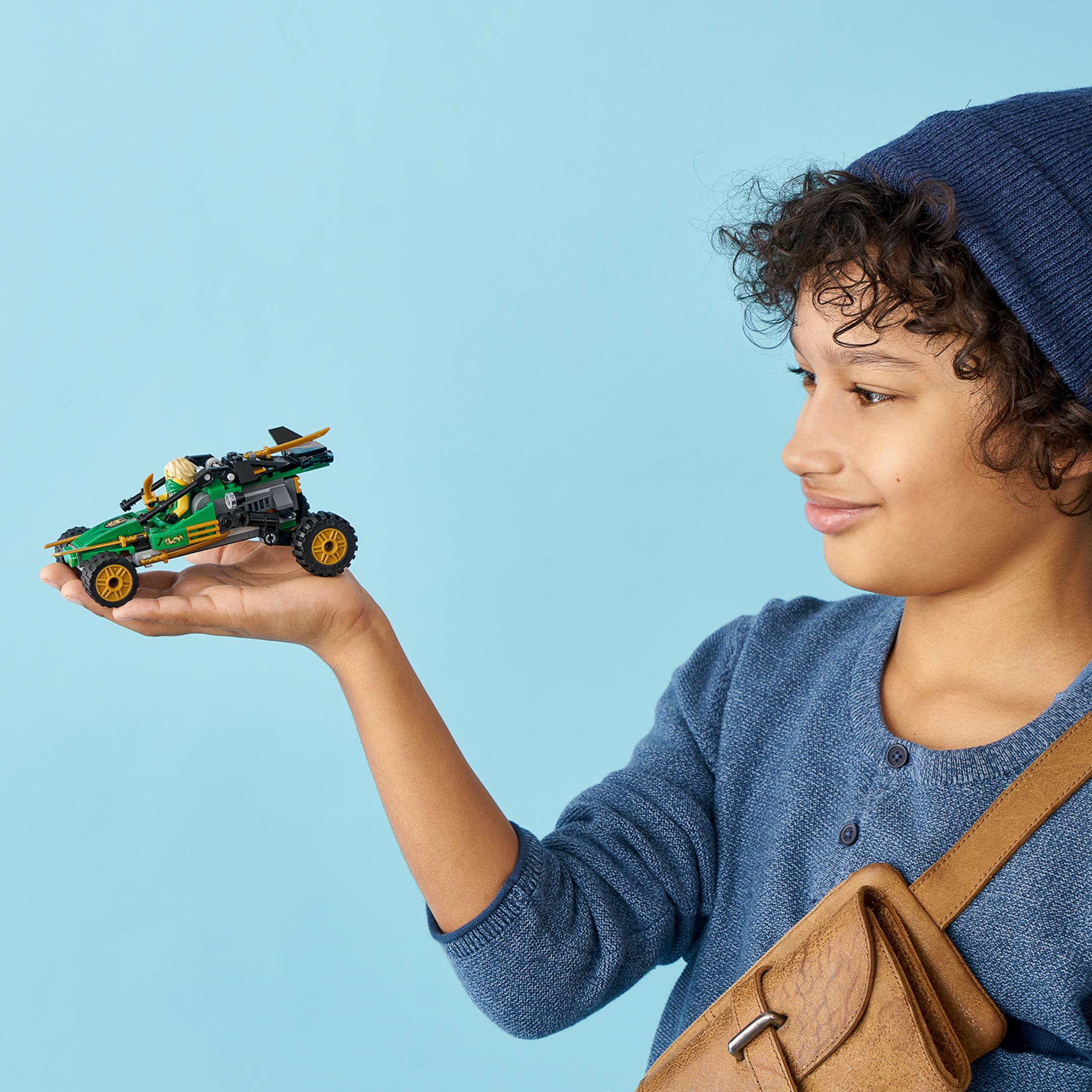 Ein Junge hält ein grünes Spielzeugauto, lächelt, trägt eine blaue Mütze und einen Pullover.
