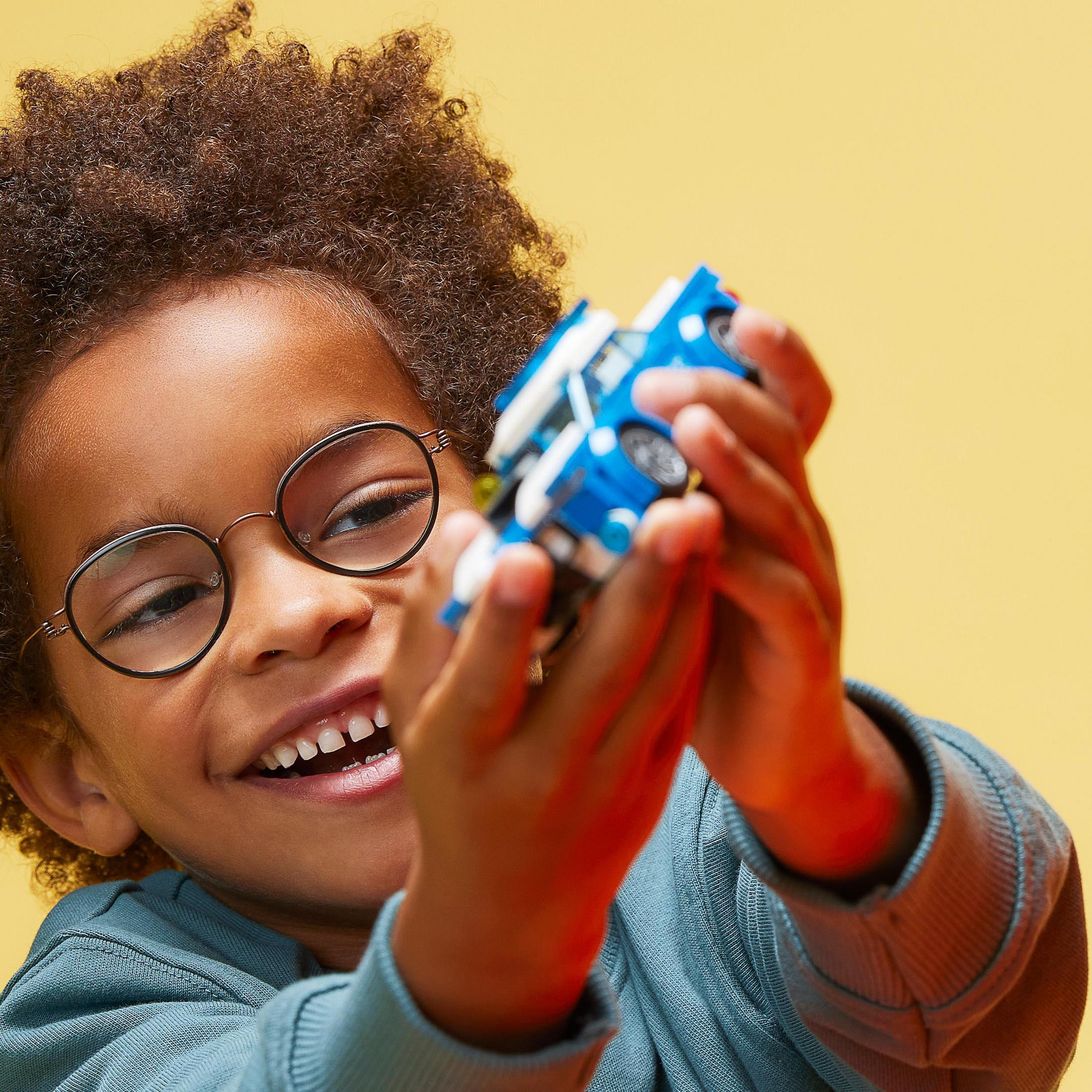 Un niño con gafas sonriendo, sosteniendo un coche de juguete azul y blanco contra un fondo amarillo.