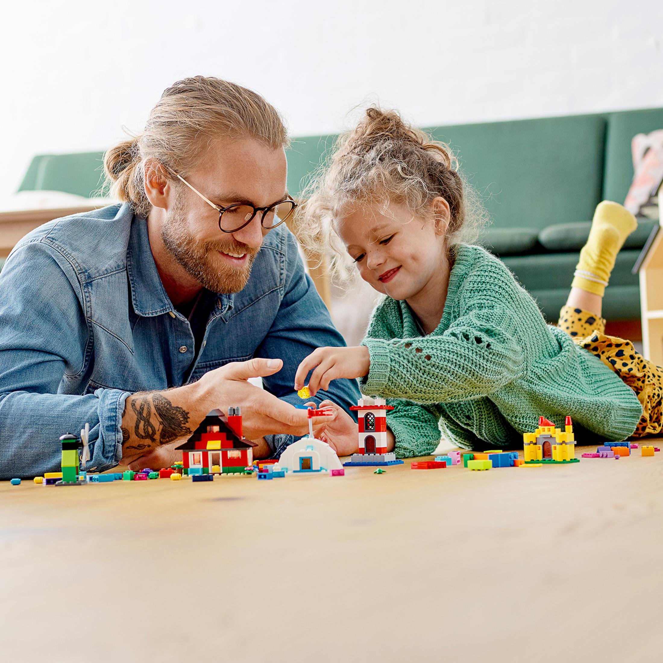 Vater und Tochter bauen mit LEGO-Steinen auf einem Holzboden. Enthält Häuser und einen Leuchtturm.