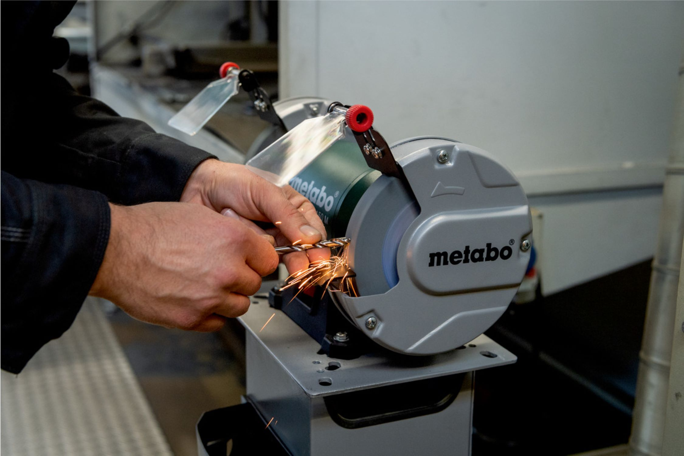 Hände eines Mannes schleifen ein Metallstück auf einer grauen Metabo-Schleifmaschine, Funken fliegen.