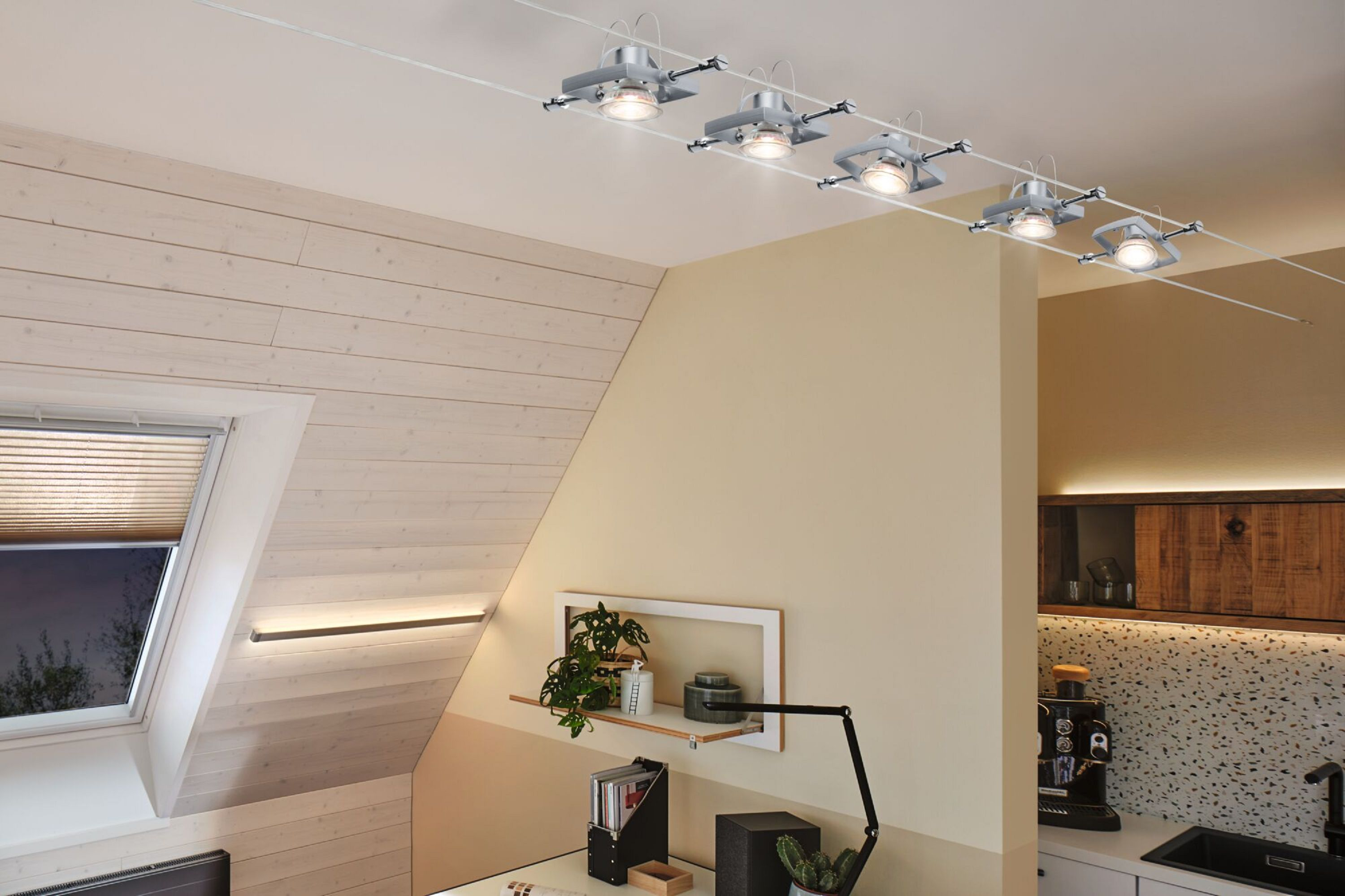 Ein Raum mit schräger Wand, Fenster und Schienenbeleuchtung. Ein Schreibtisch mit Lampe befindet sich im Vordergrund.