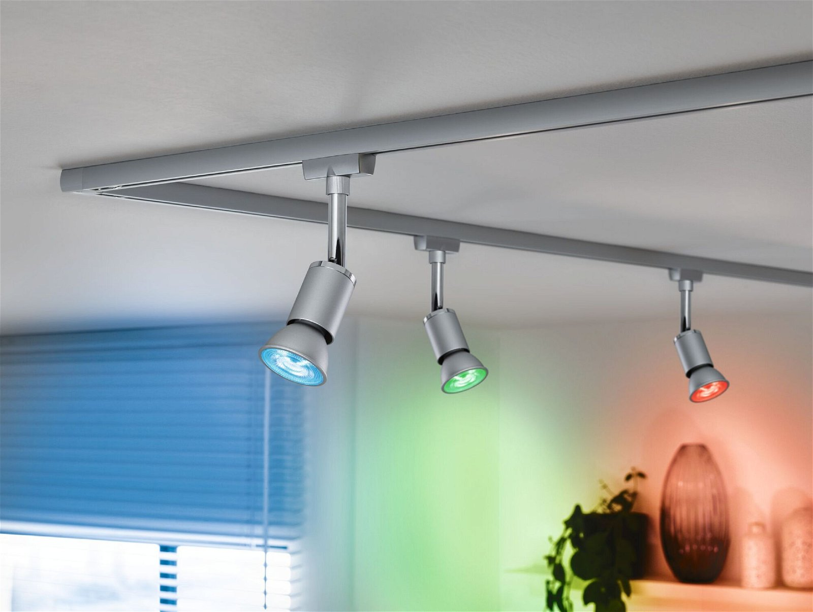 Drei silberne Schienenstrahler mit farbigem Licht, das auf eine Wand scheint. Ein Licht scheint blau, grün und rot.